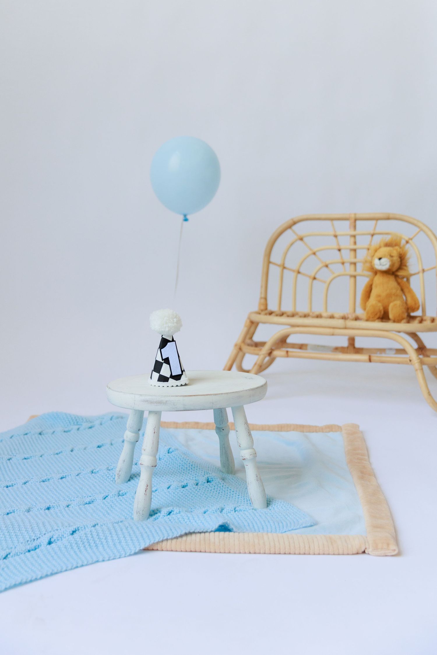 Minimalist nursery scene with a light blue balloon, teddy bear, and white stool on a knitted rug.