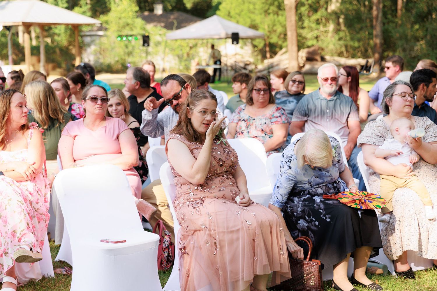 Guests at an outdoor garden party celebration wearing pink and white attire mingle and toast under trees.