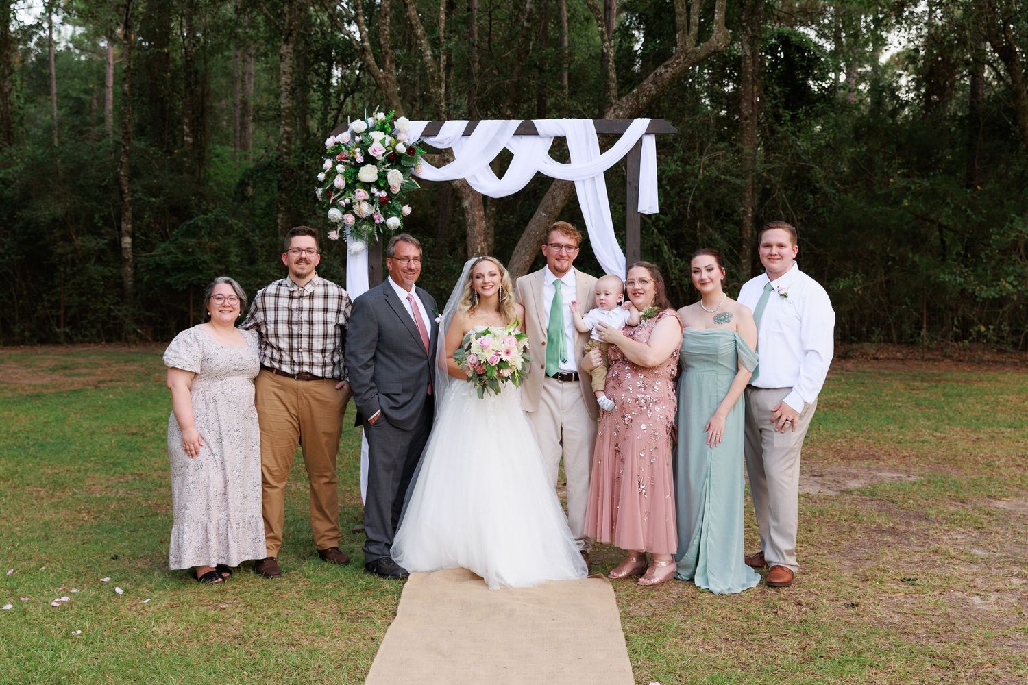 Wedding party poses together outdoors in a natural wooded setting with soft pastel colors and casual attire.