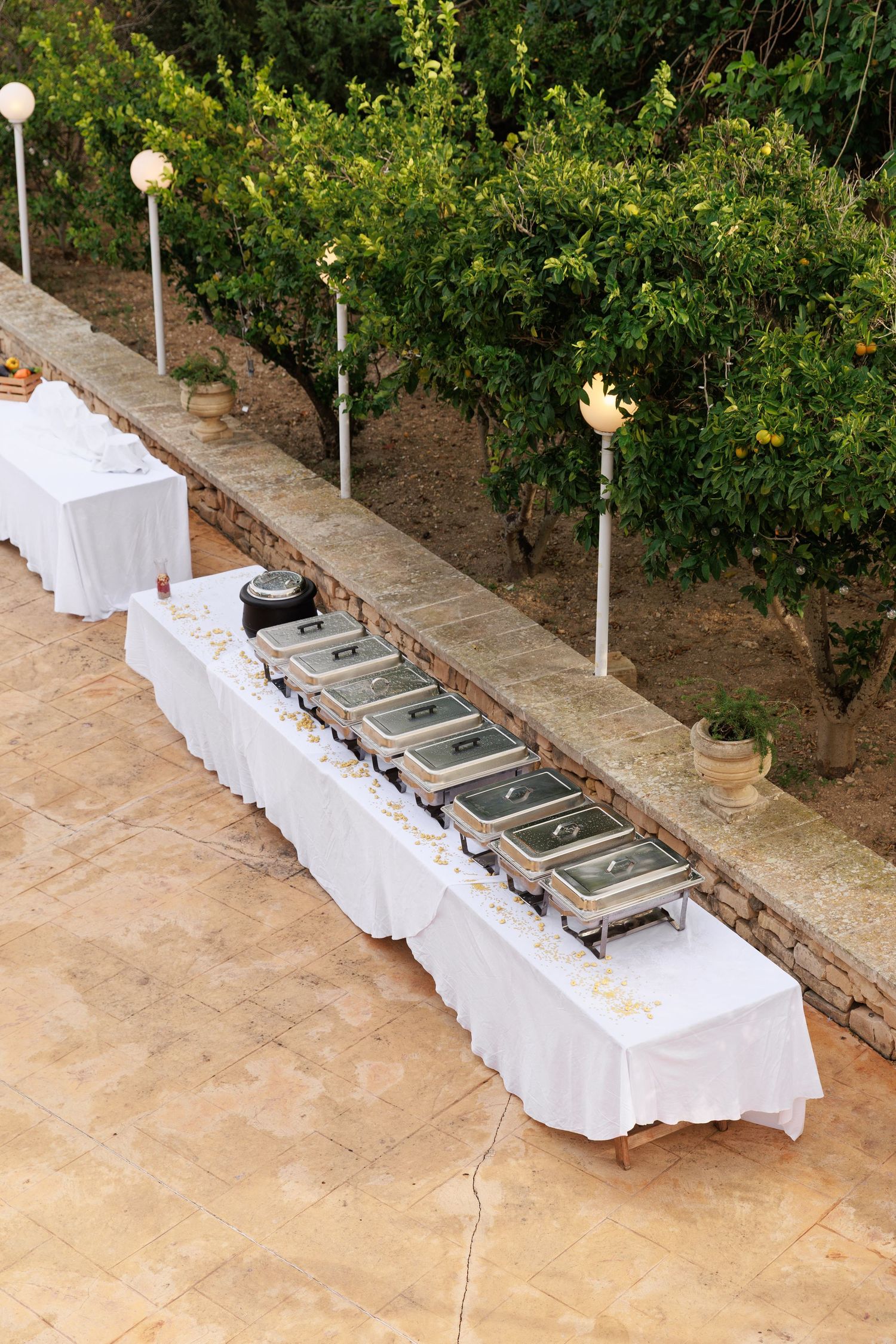 Outdoor buffet setup with white tablecloths and silver chafing dishes arranged along a path bordered by citrus trees.