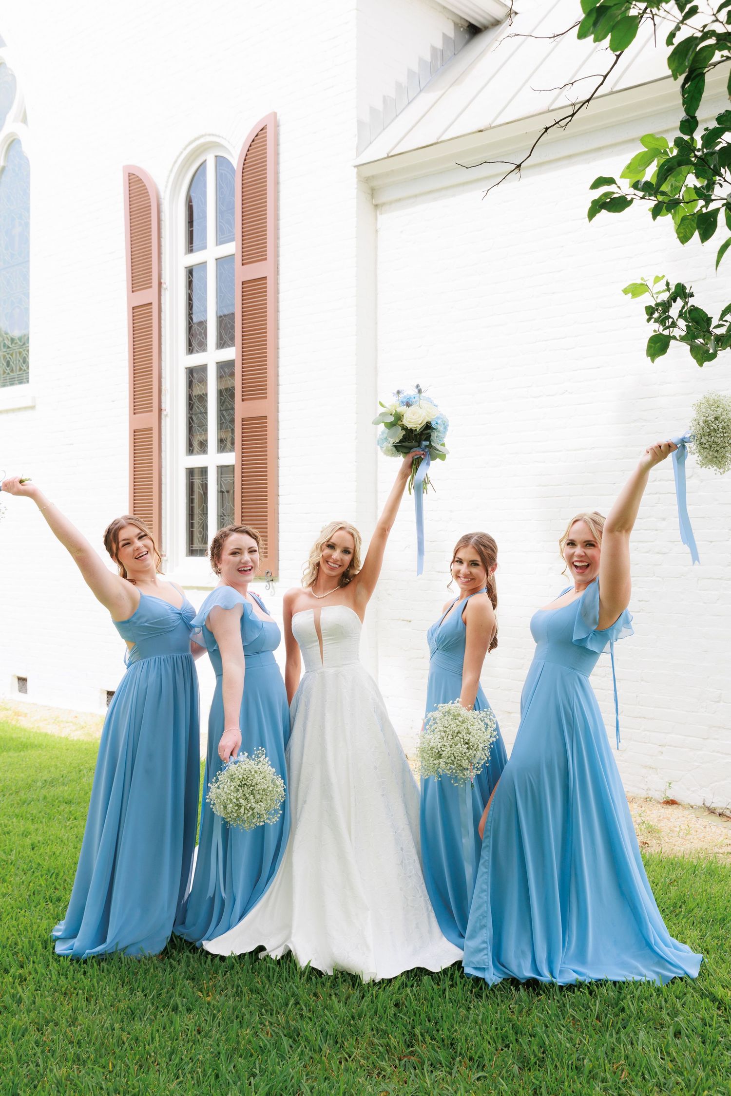 Wedding party in blue bridesmaid dresses pose joyfully with bride outside church.