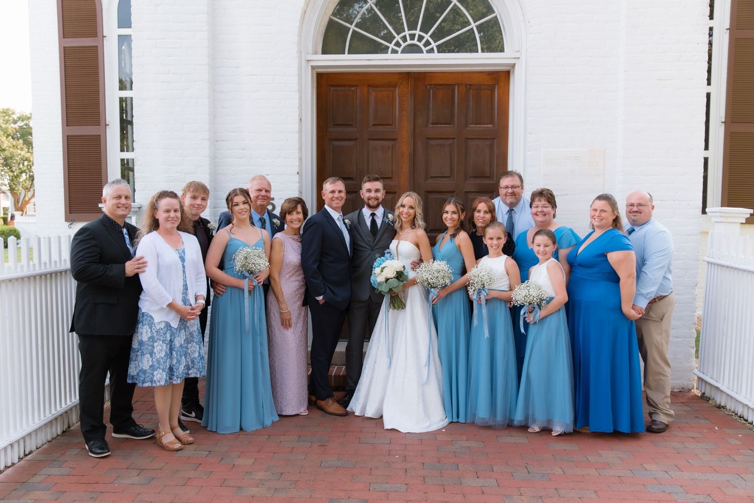 Wedding party in coordinated blue and formal attire poses together on church steps.