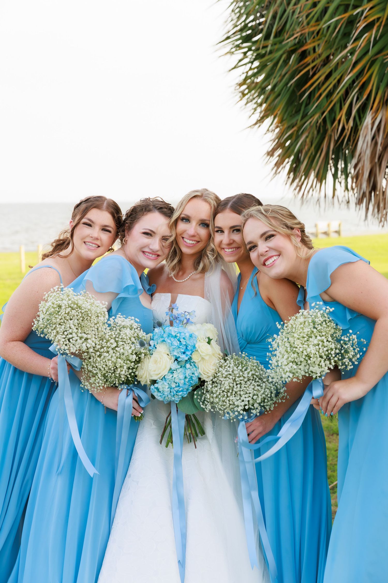 A bridal party in light blue dresses holding bouquets poses outdoors under a palm tree.