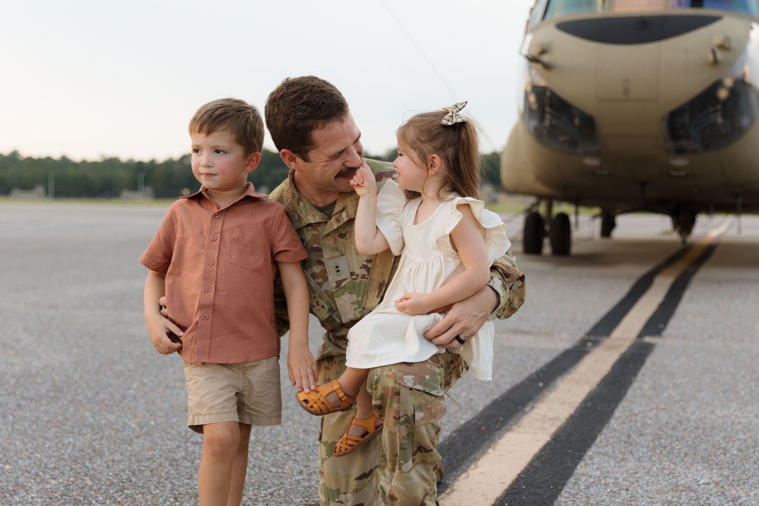 A service member in camouflage uniform holds two children on a tarmac with a military aircraft in the background.