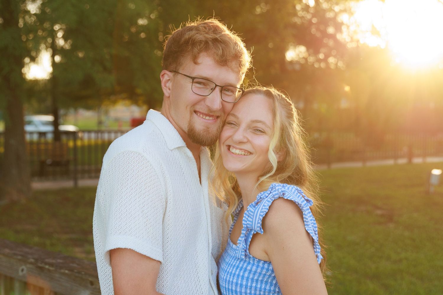 Happy couple smiling together outdoors at golden hour with warm sunset light filtering through green trees.