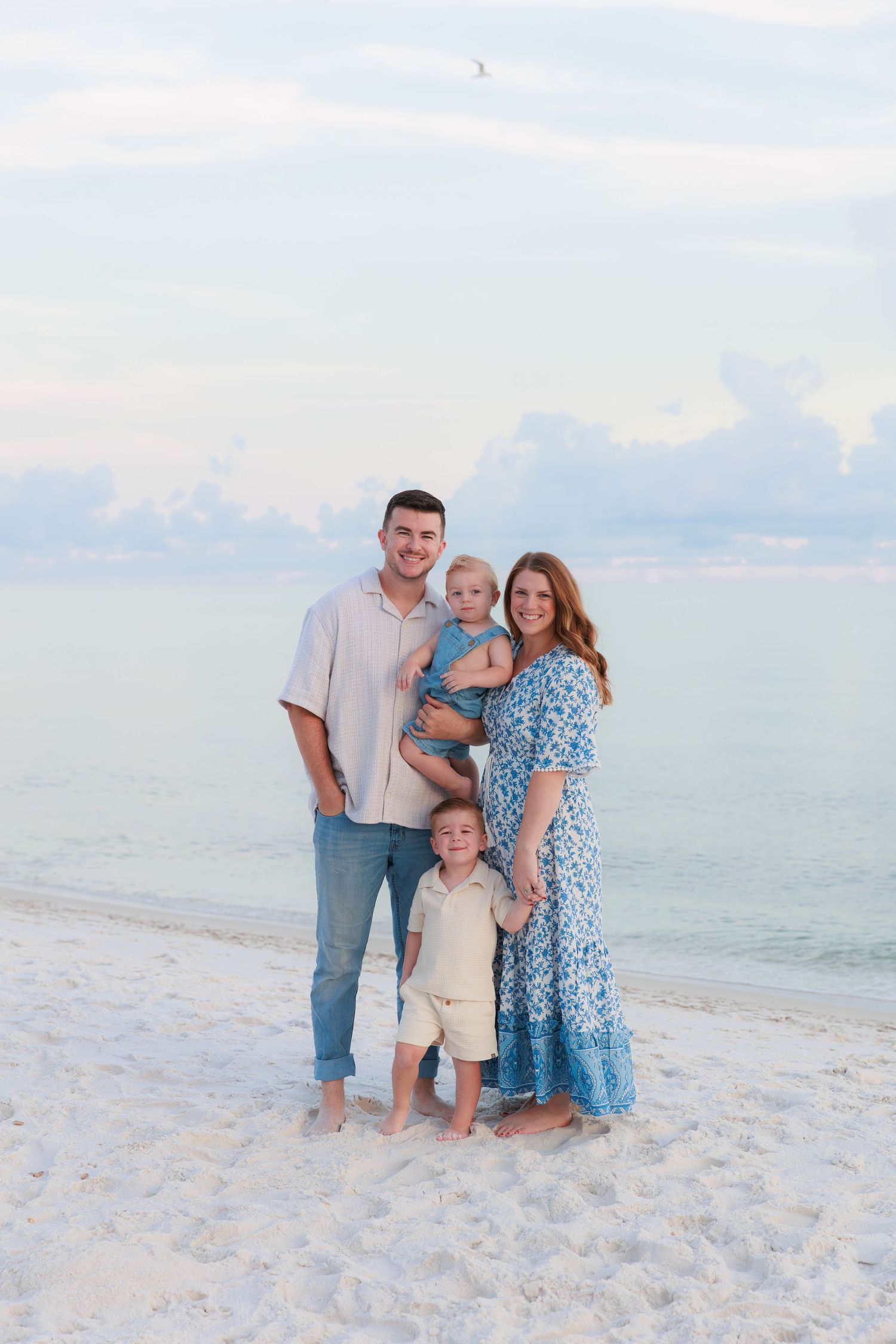 Happy family of four posing on white sandy beach with calm ocean and cloudy sky backdrop.