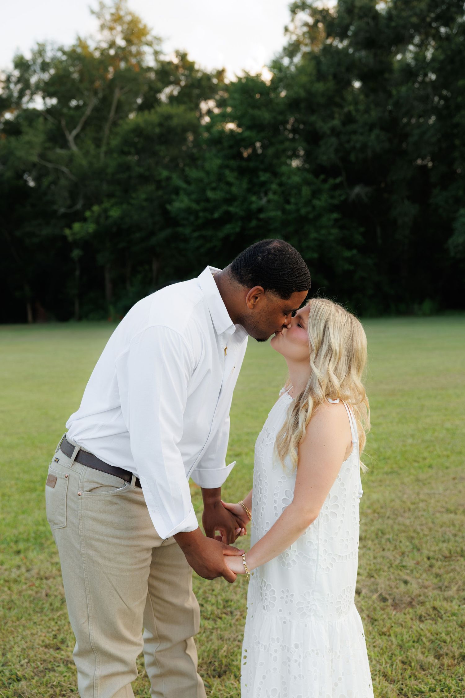 A couple shares a romantic kiss in a sunlit green field, the man in white shirt and the woman in a white lace dress.
