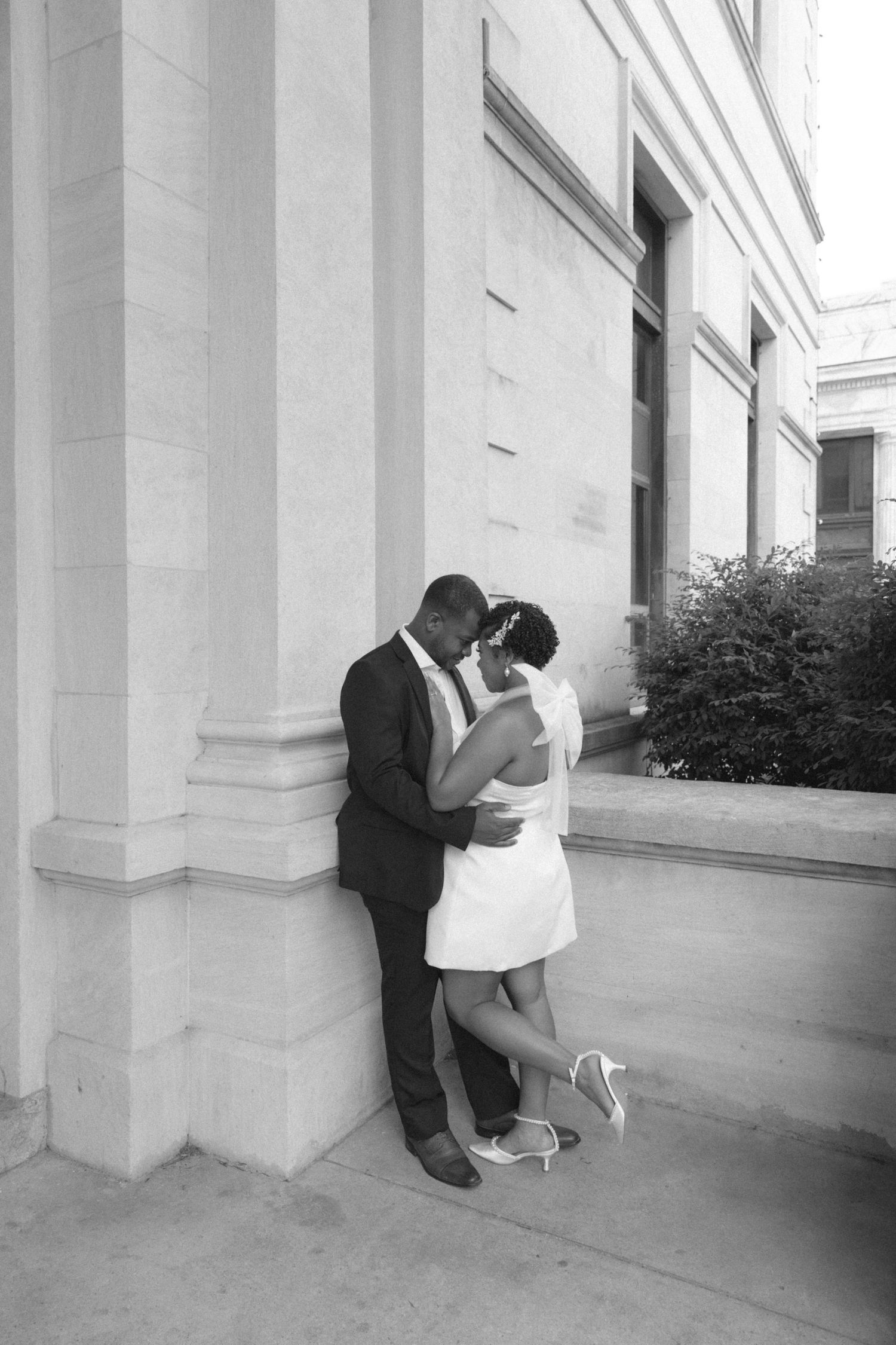 Black and white photo of a couple sharing an intimate embrace against a stone column outdoors.