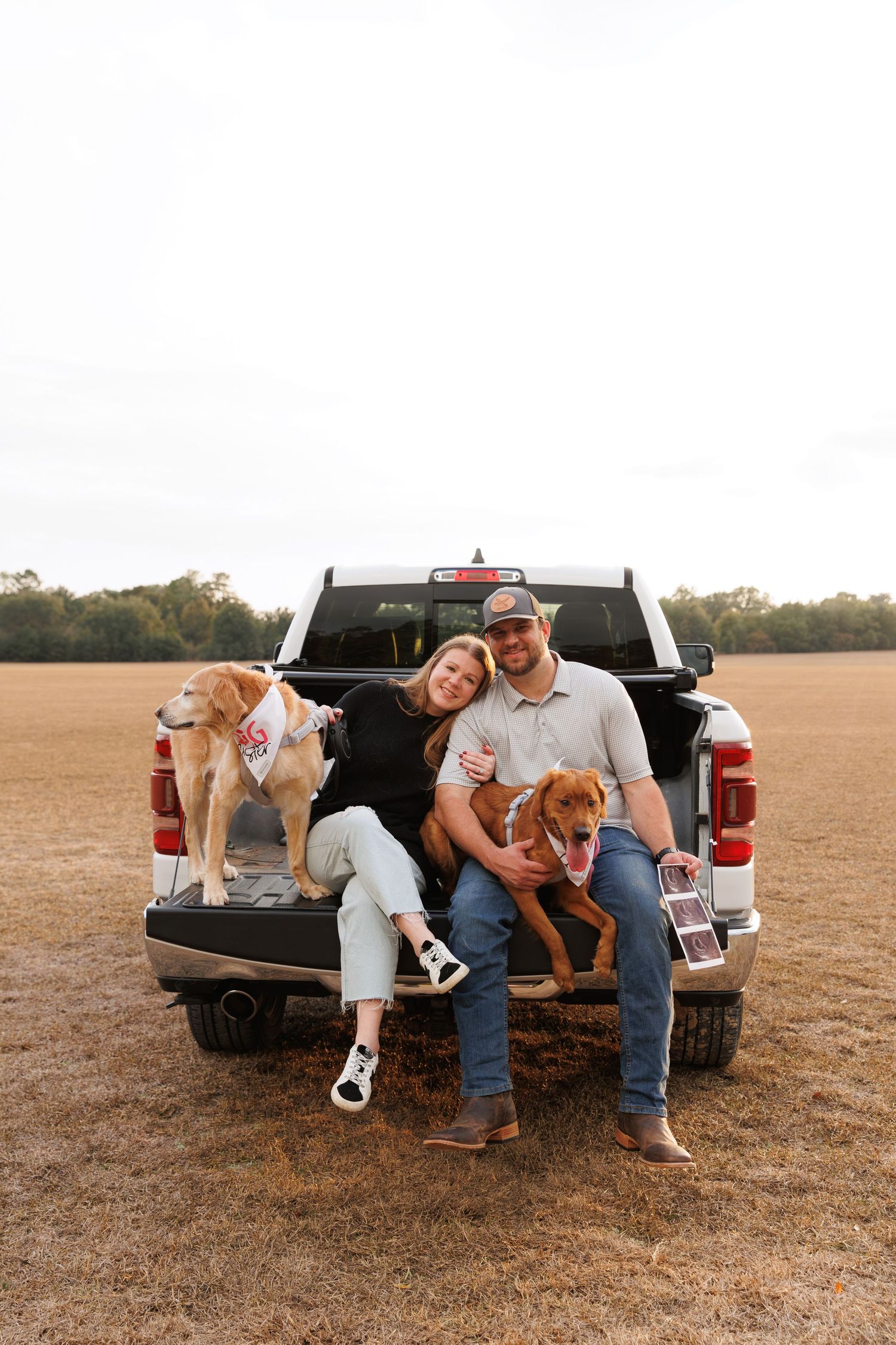A couple sits with their two dogs on the tailgate of a pickup truck in a brown field.