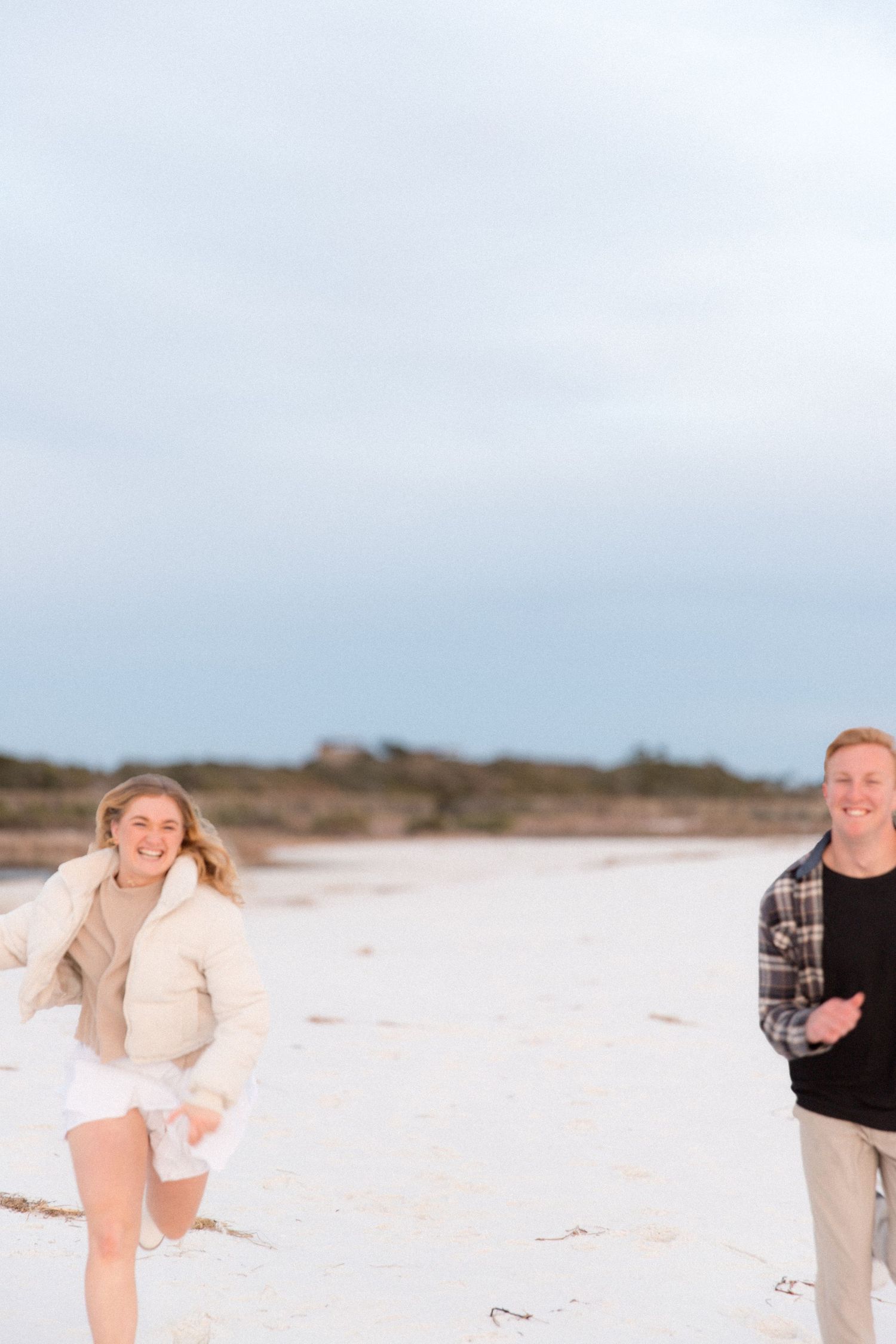 A playful couple runs and laughs together on a bright sandy beach on an overcast day.