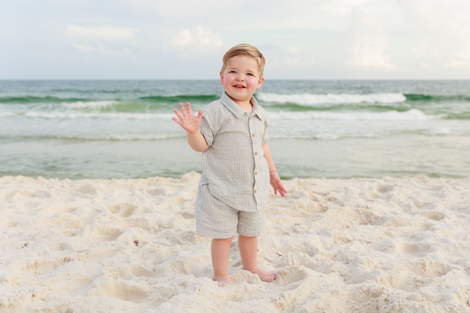 A child wearing gray romper plays and walks along a sandy beach with ocean waves in the background.