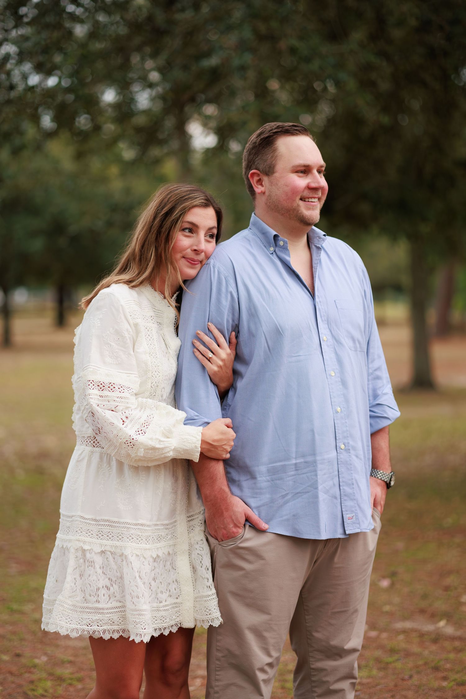 A couple in a park setting with the woman wearing a white lace dress and the man in a light blue button-down shirt.