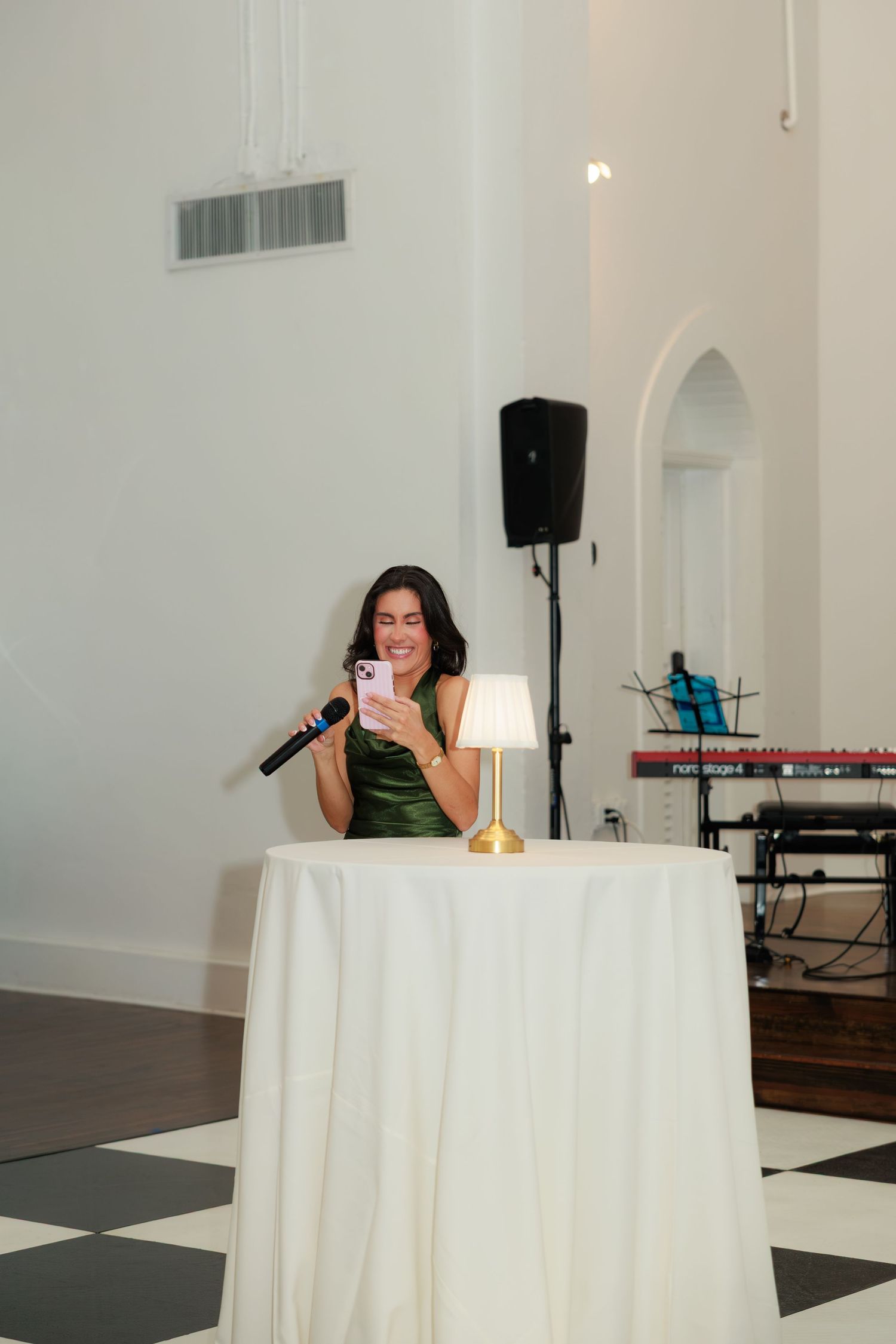 Woman in green dress smiling at phone while holding microphone at elegant wedding reception.