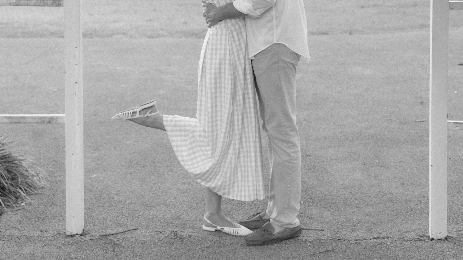 Black and white photo of a couple embracing outdoors, woman in plaid skirt lifting one leg romantically.
