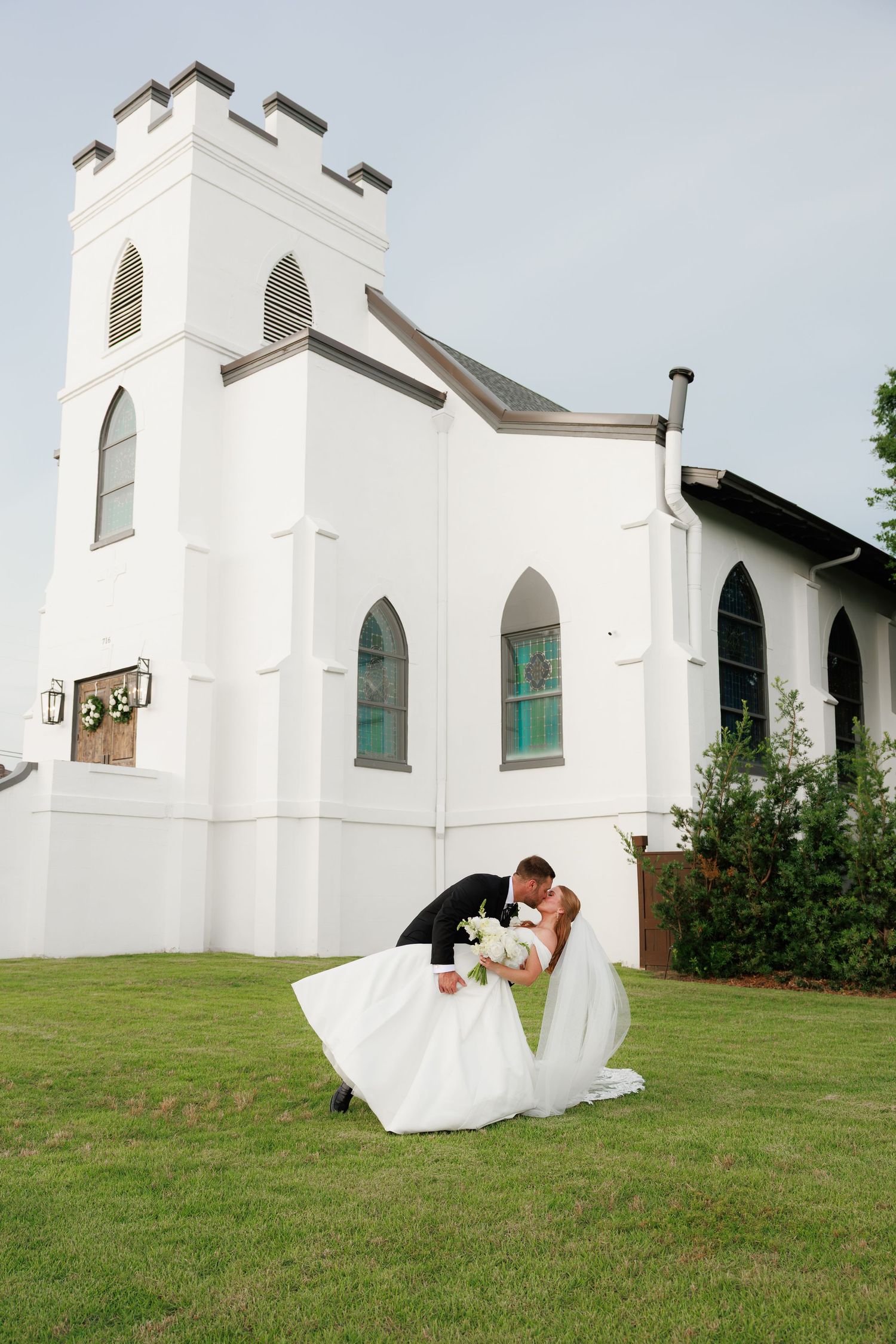 Bride and groom share a romantic dip kiss on green lawn outside a white historic church on their wedding day.
