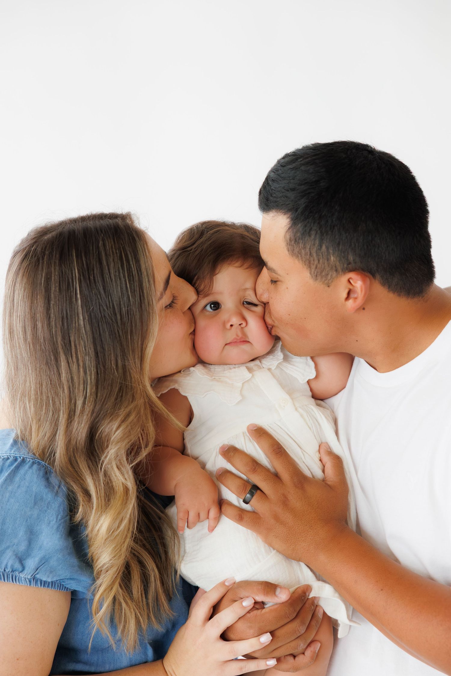 Parents kissing their baby on the cheeks in a sweet family portrait against a white background.
