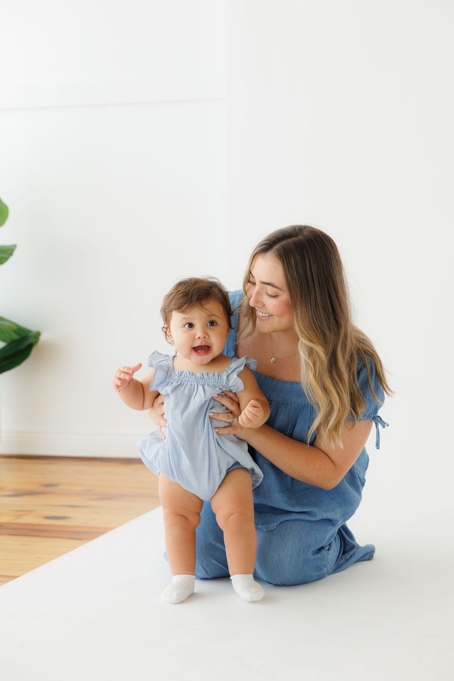 A smiling mother in a blue dress sits on the floor holding her happy baby girl in a matching blue romper.