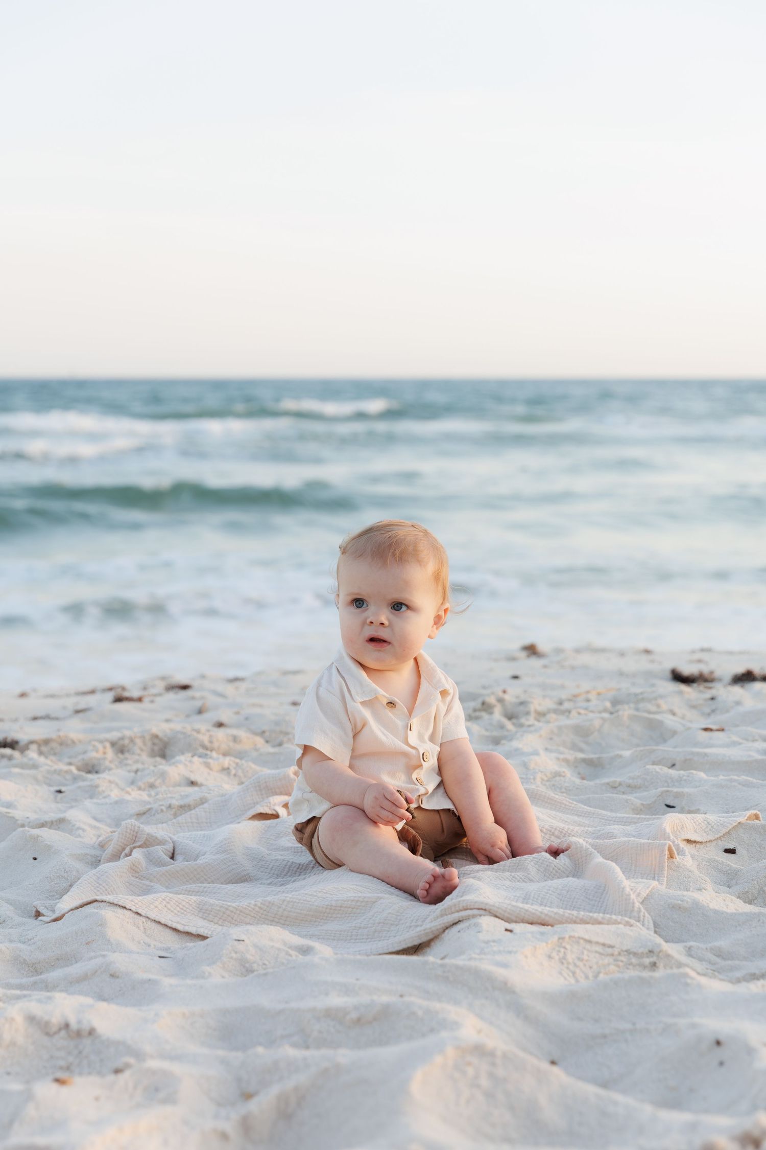 A cute baby sits on a white blanket on a sandy beach with ocean waves in the background.