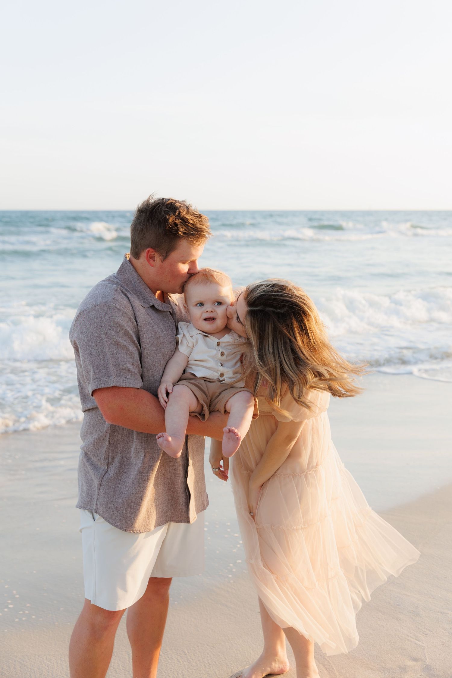 A young family with a baby enjoys a sunny beach day, with parents kissing their infant by ocean waves.