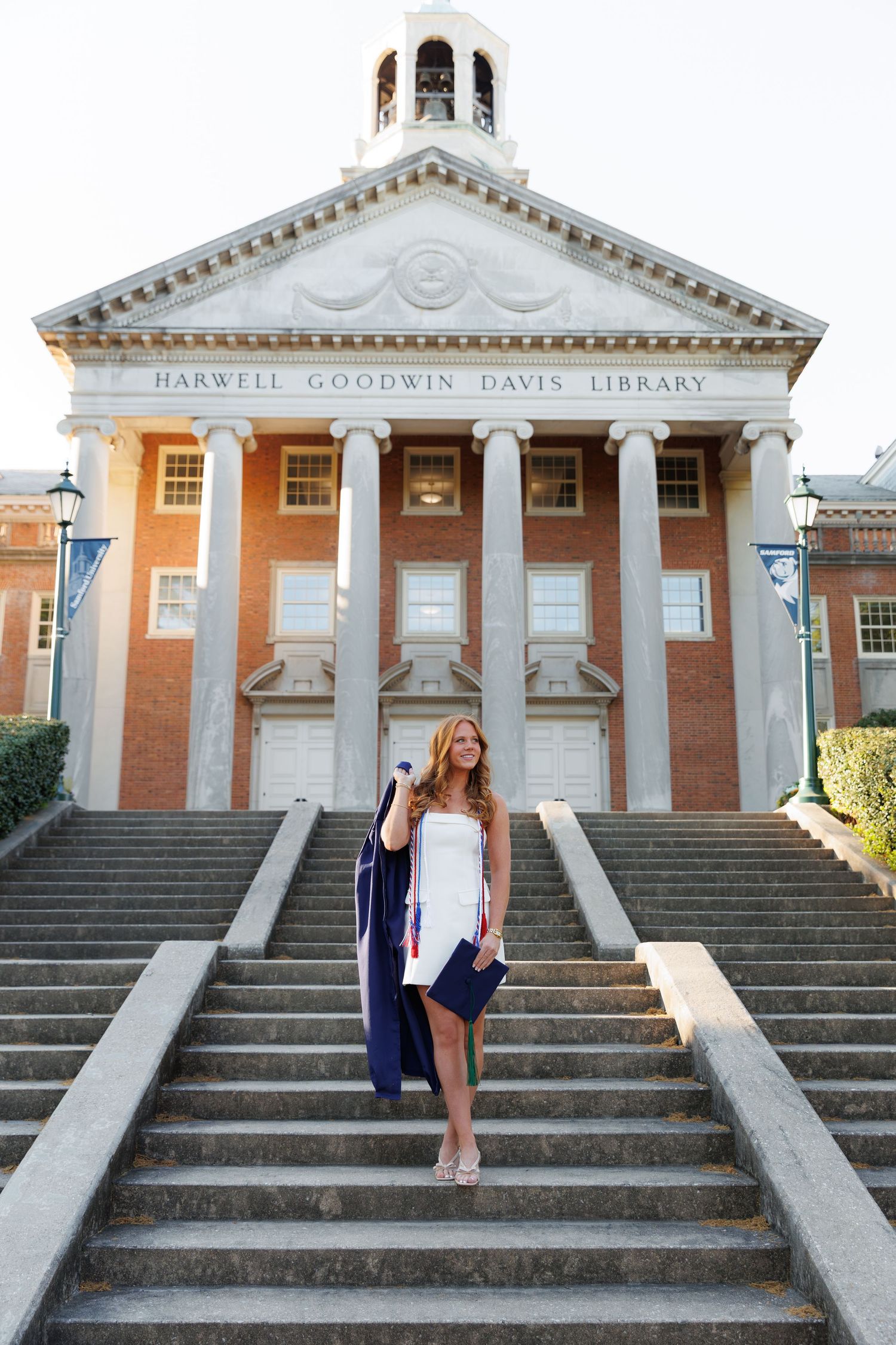 A graduate in white dress holds cap and gown on steps of Harwell Goodwin Davis Library brick building.