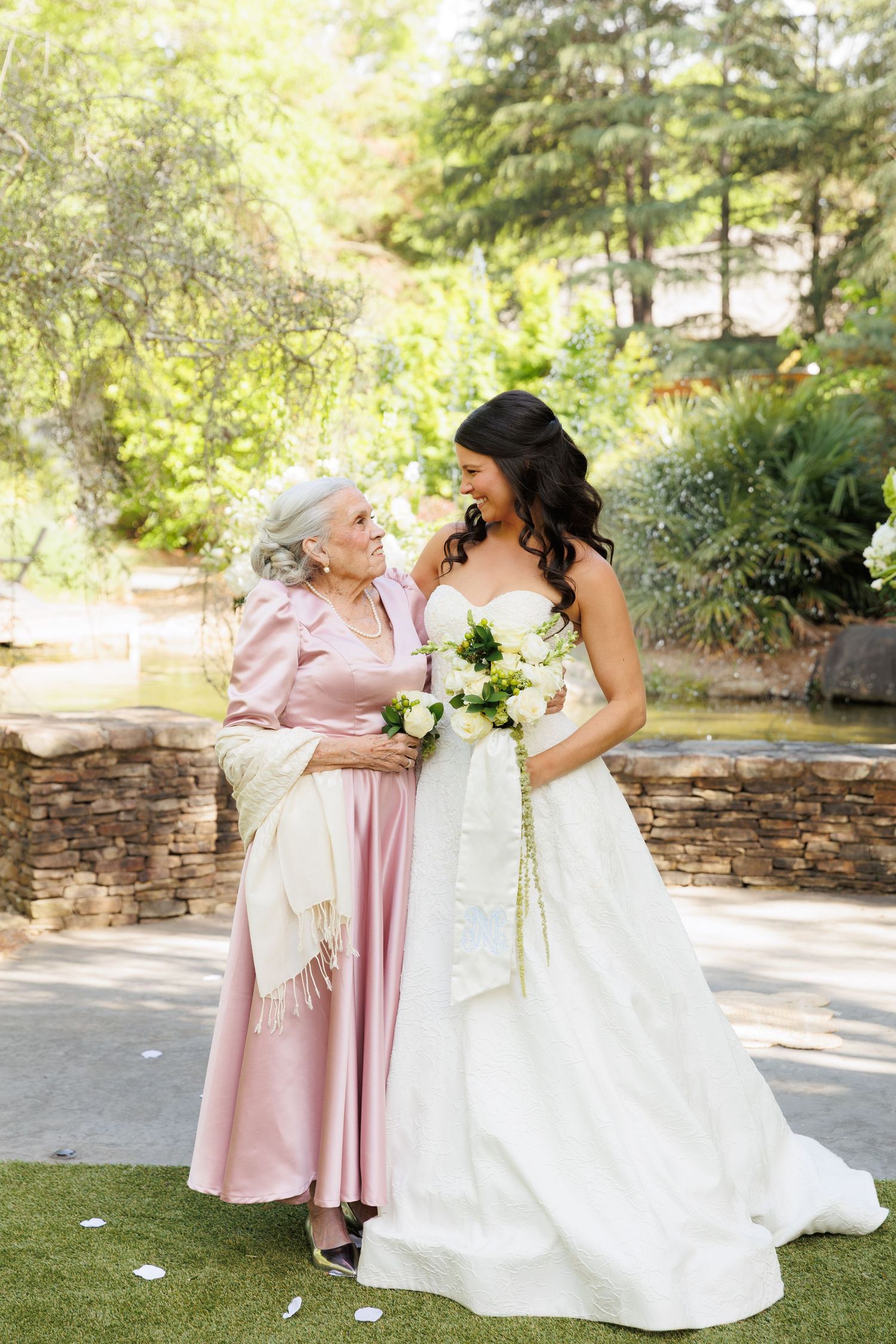 Bride in white gown holding bouquet stands with elderly woman in pink dress outdoors at garden wedding venue.