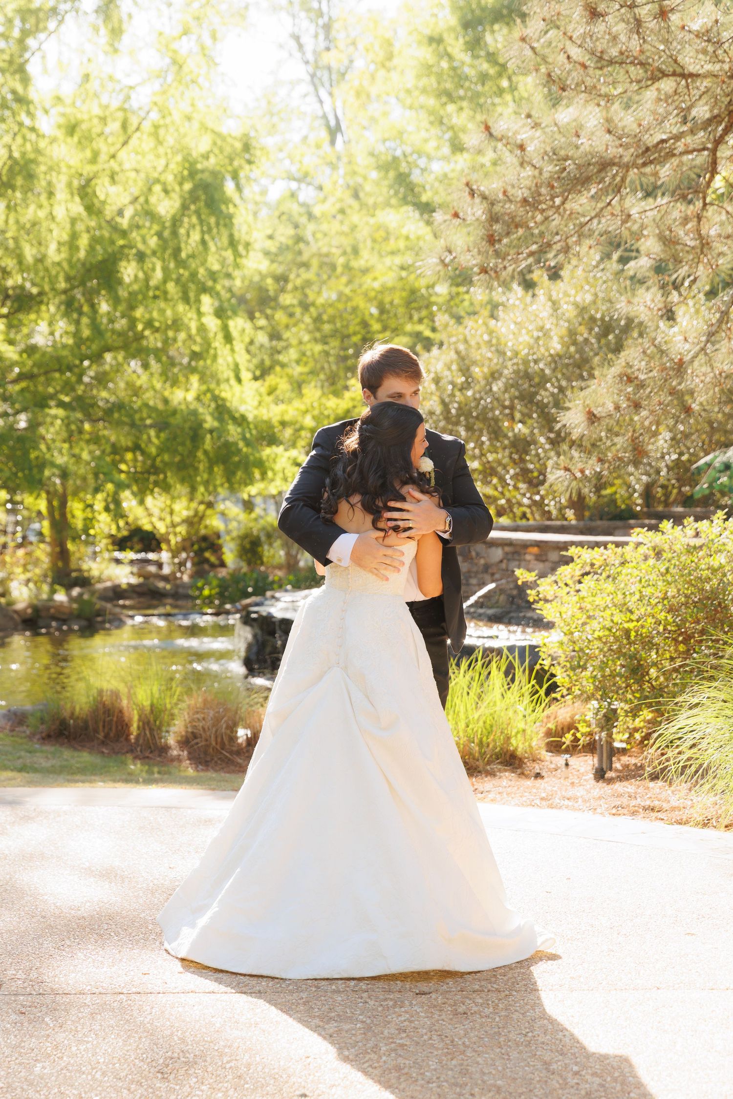 Bride and groom sharing a romantic embrace in a sunlit garden with a peaceful pond in the background.