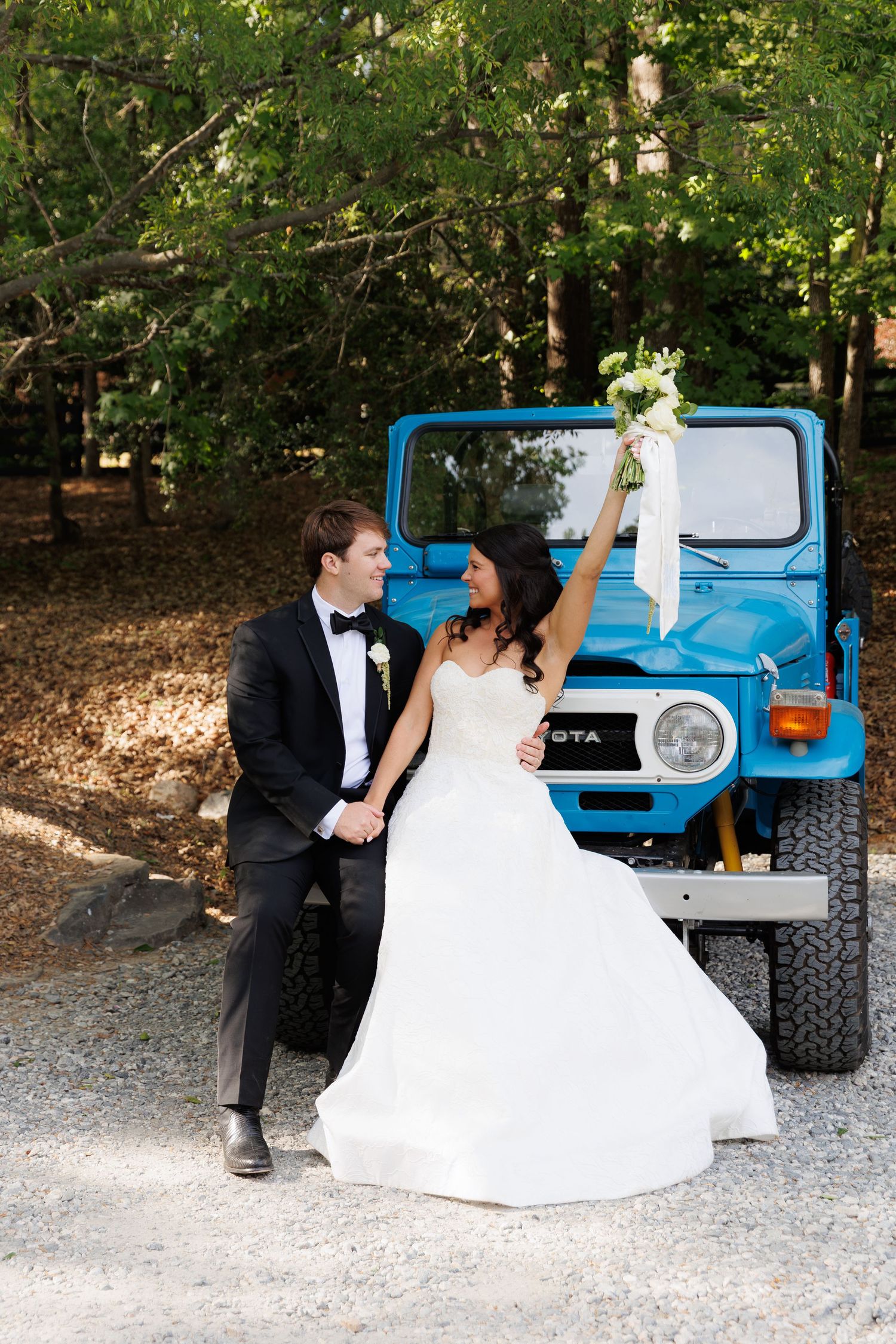Bride and groom pose in front of a vintage blue Toyota FJ40, bride raising bouquet joyfully.