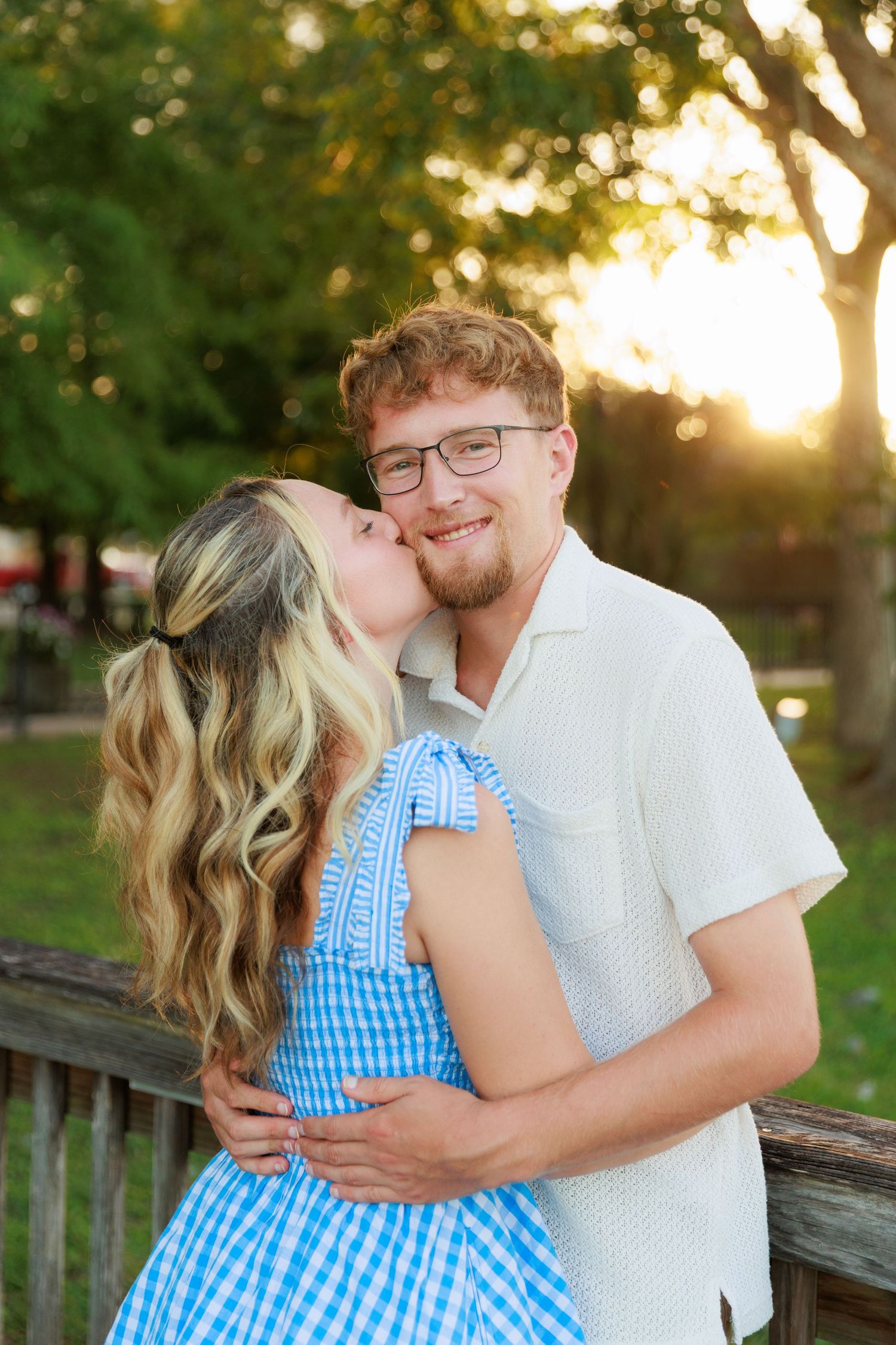 A woman in a blue gingham dress kisses a smiling man in glasses outdoors at golden hour.