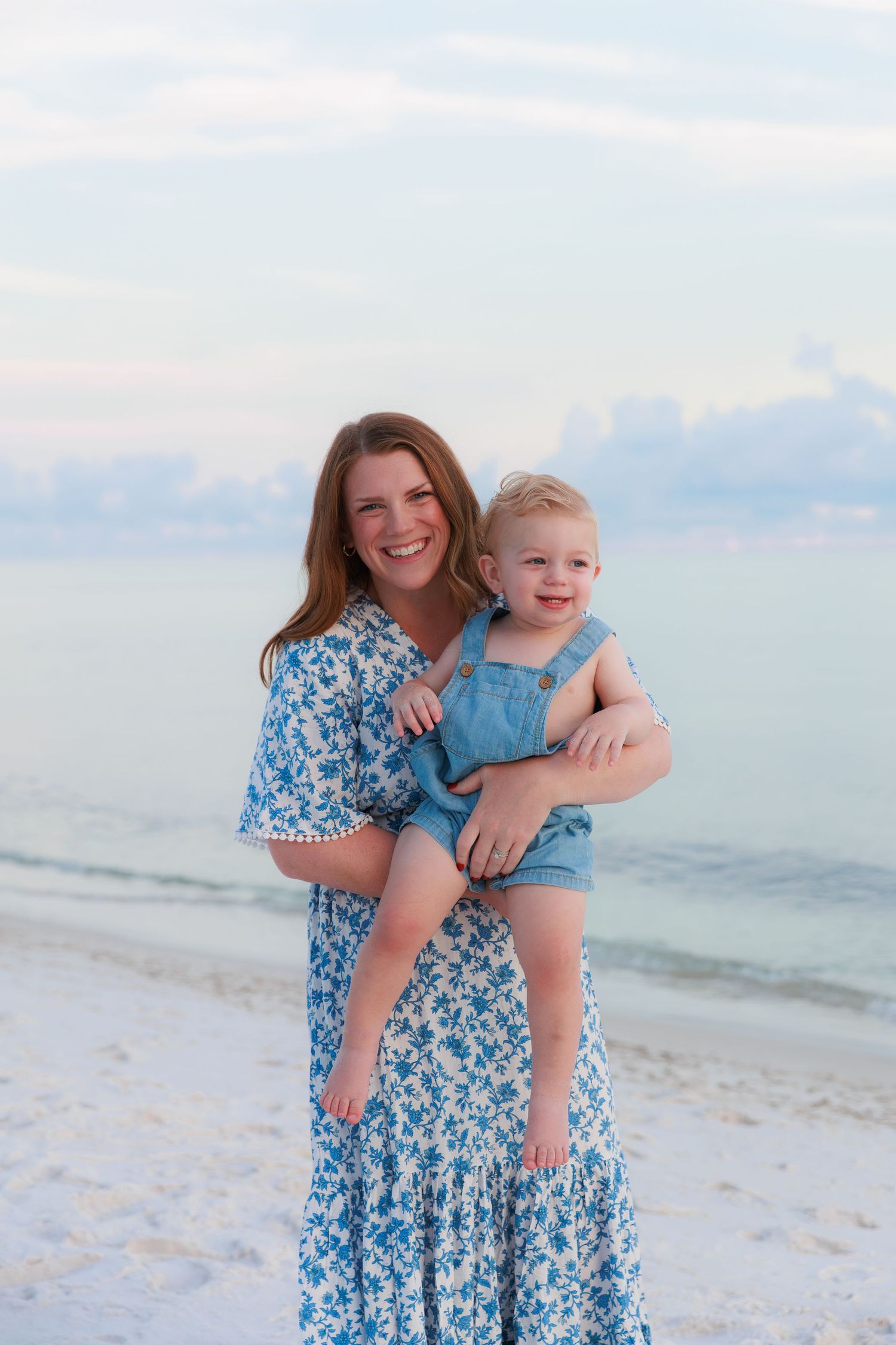 Smiling mother in blue floral dress holds toddler boy in denim overalls on a white sand beach at sunset.