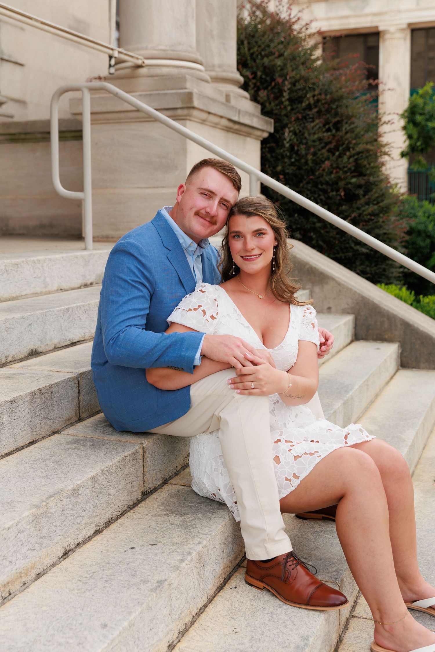 Couple sitting on stone steps, man in blue blazer embracing woman in white lace dress, smiling at camera.