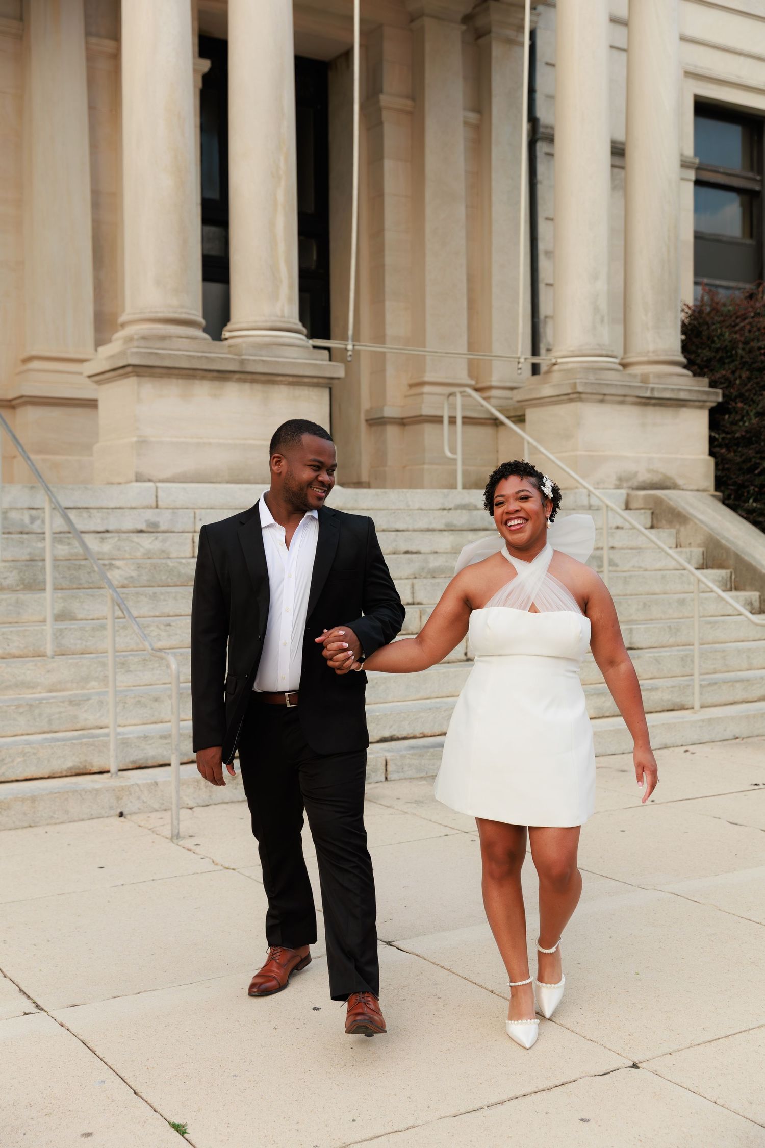 A joyful couple walks hand-in-hand outside a grand building, dressed in wedding attire, smiling at each other.
