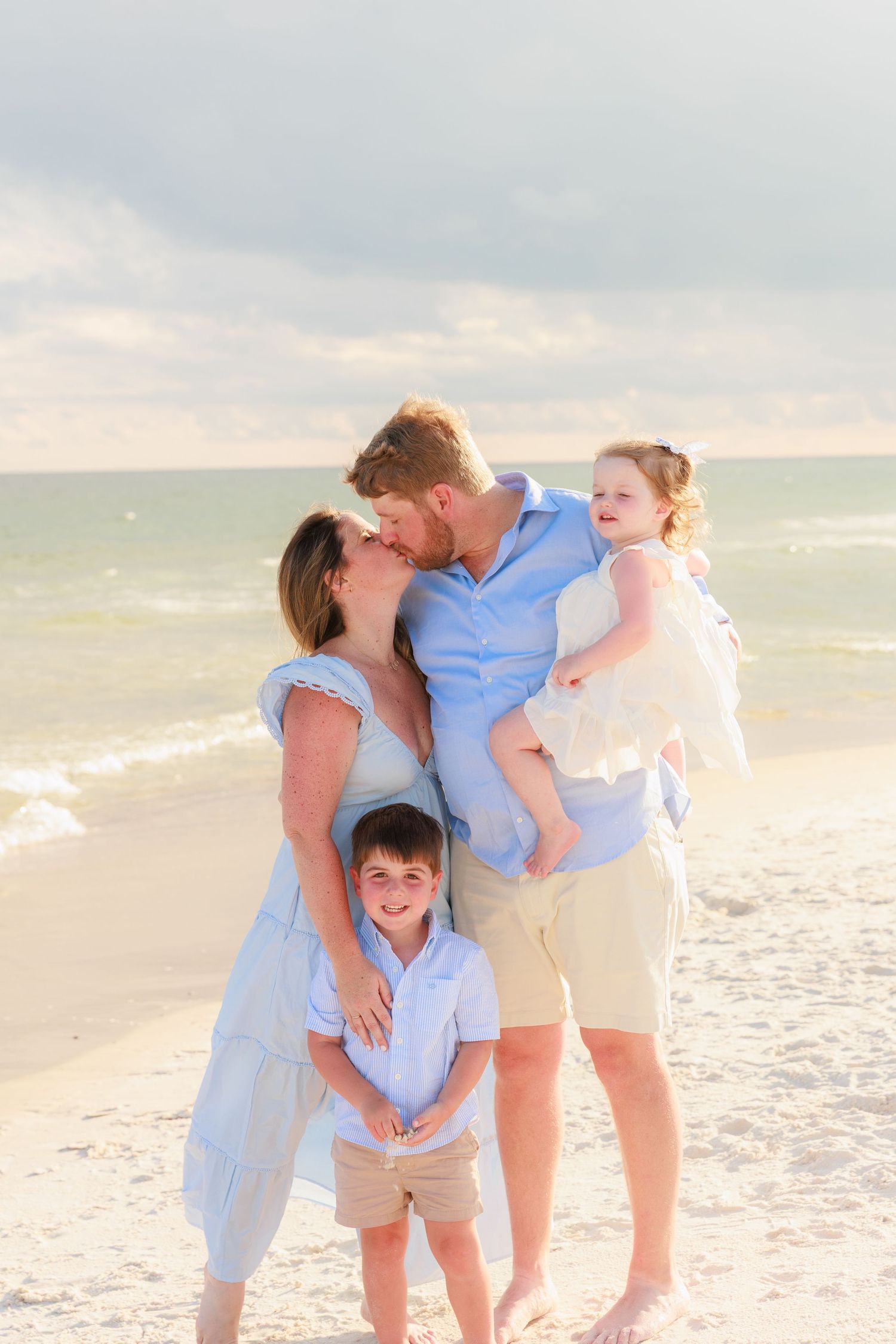 Family of four posing on a sunny beach, parents sharing a kiss while holding young children dressed in coordinating blue outfits.