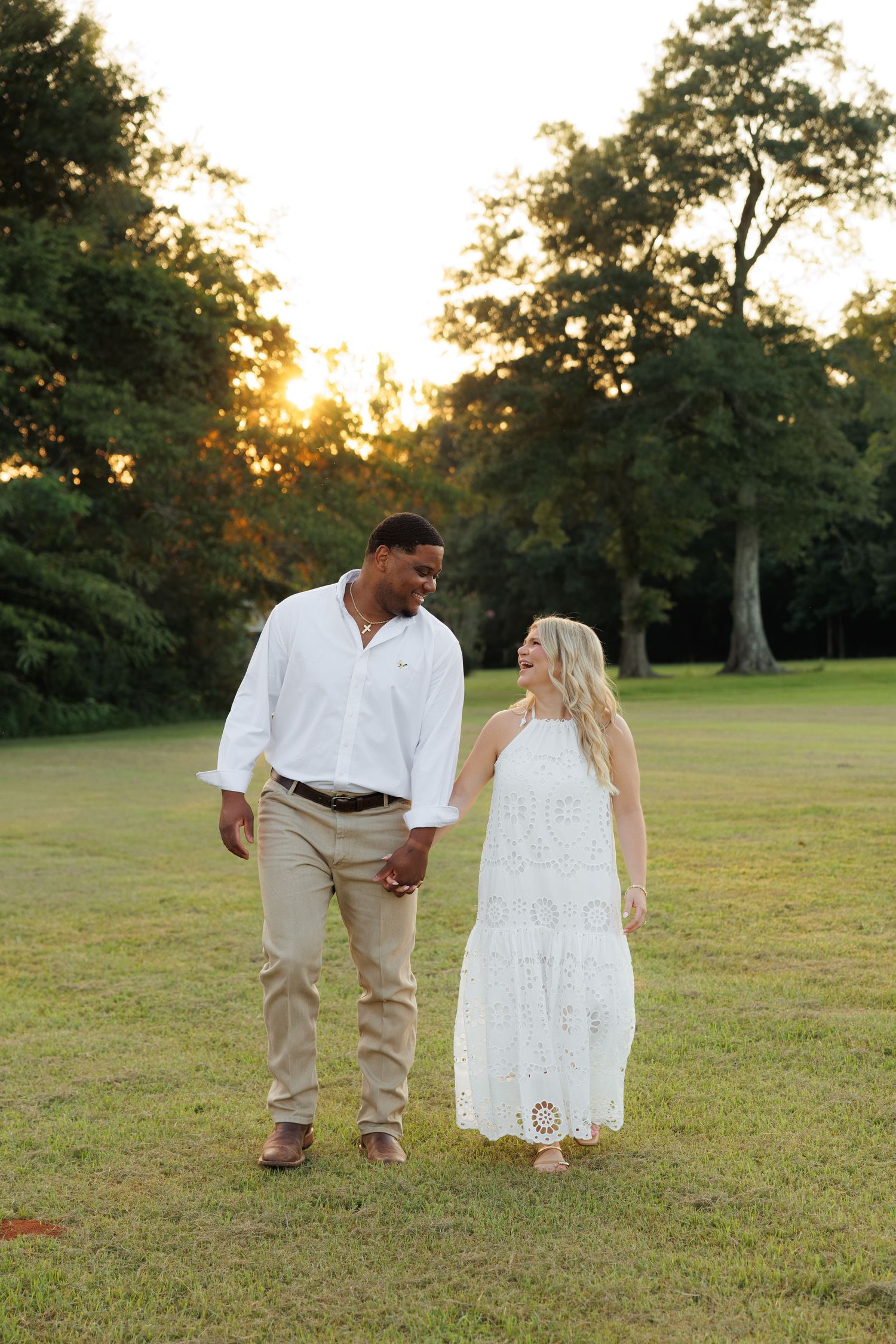 Couple holding hands walking on green grass at sunset, man in white shirt and khakis, woman in white lace dress.