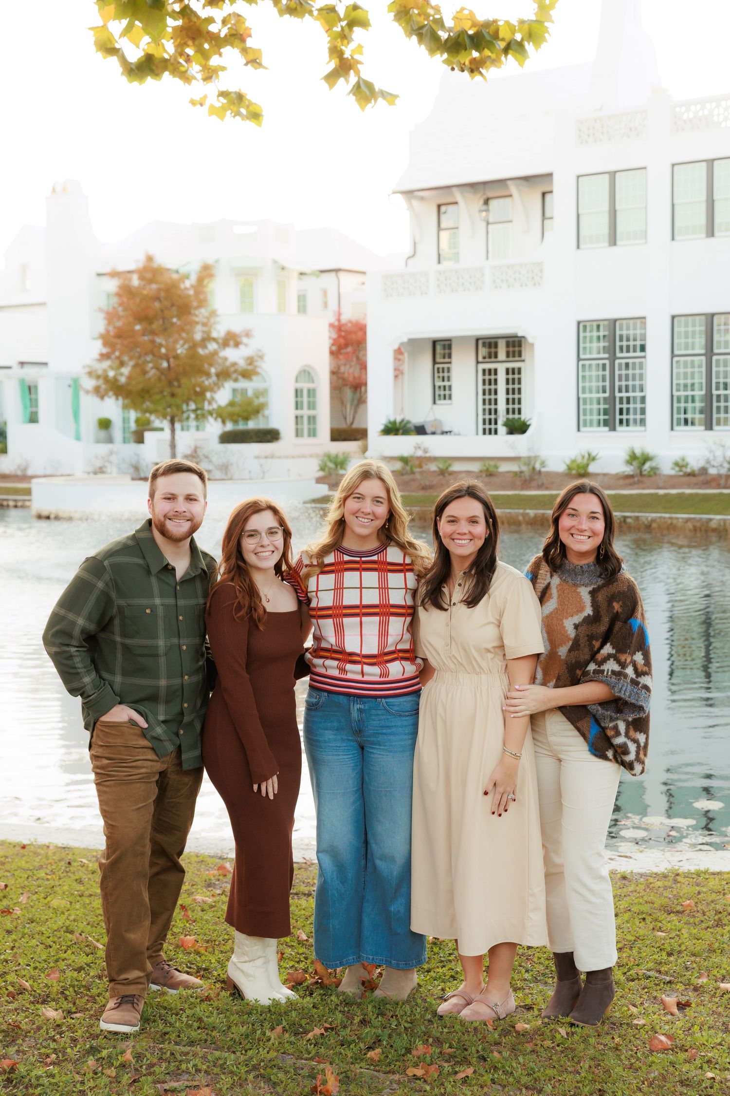 A group poses together outdoors on grass near a white building and pond on an autumn day.