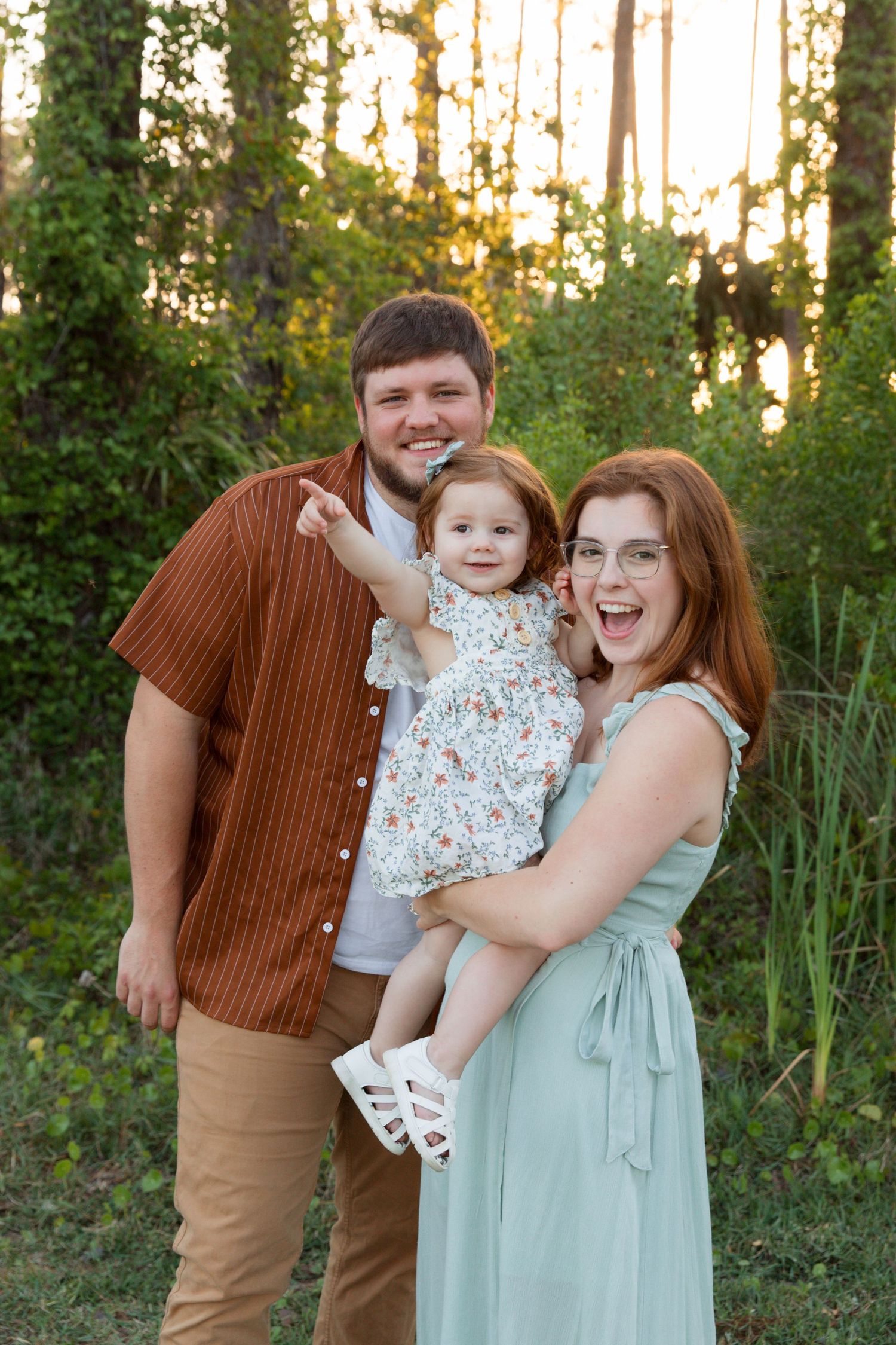 A family poses together outdoors in coordinated outfits against a backdrop of trees at sunset.