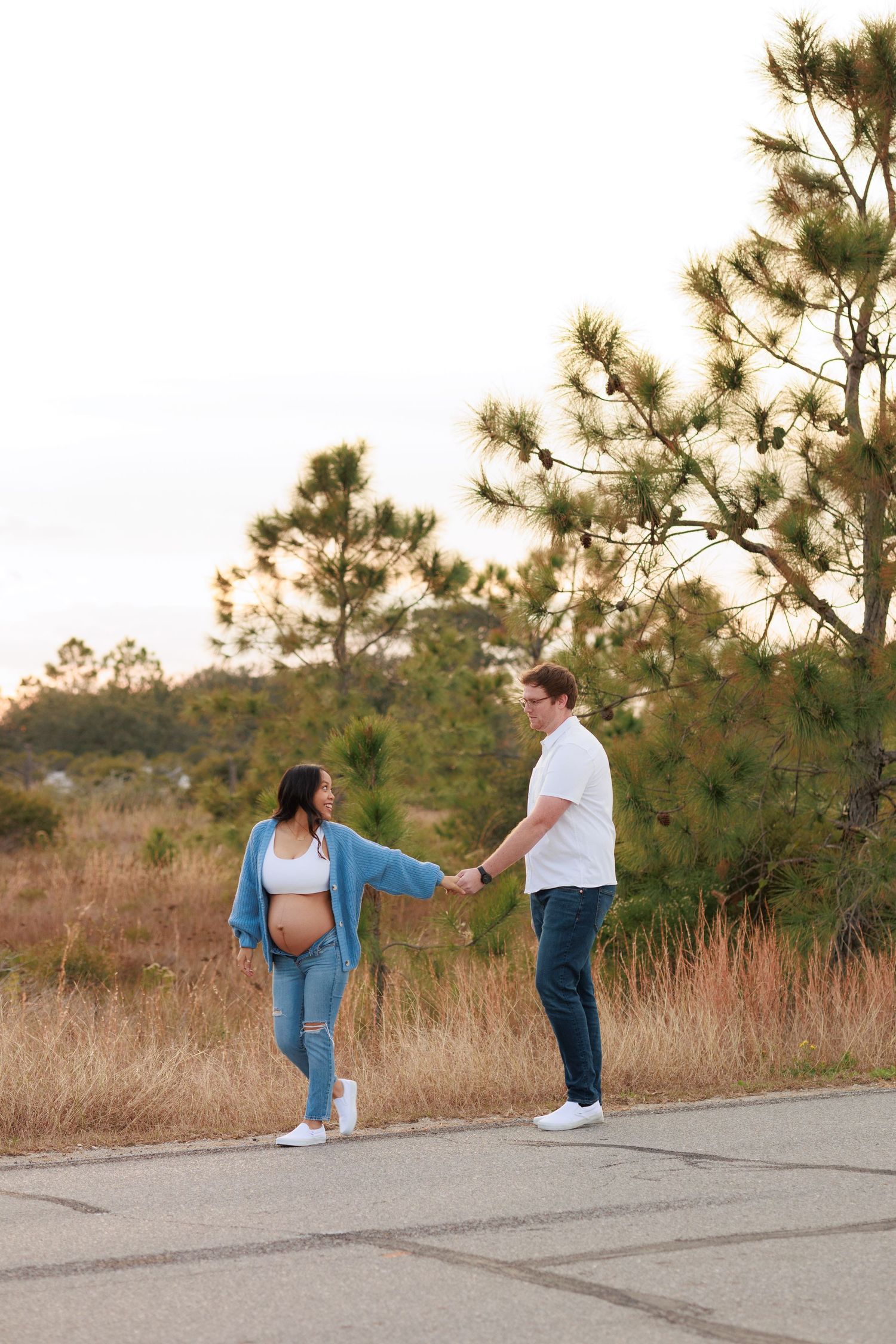 A couple holds hands and spins together on a paved path surrounded by tall grass and pine trees at sunset.