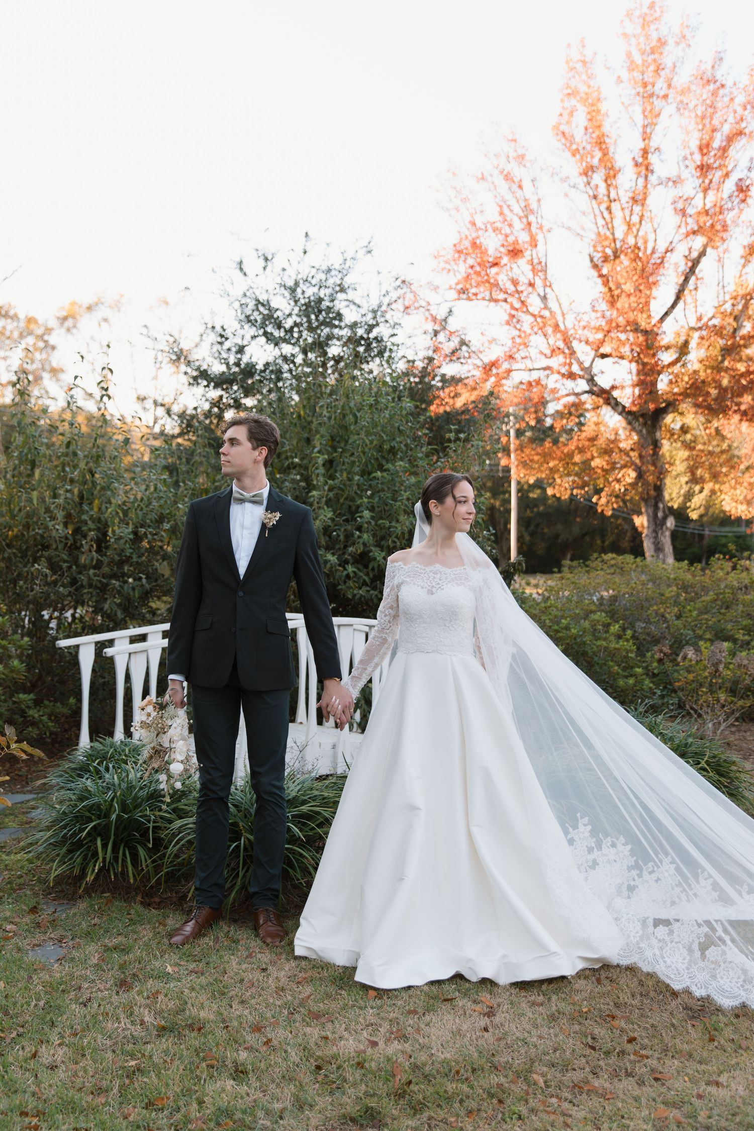 A romantic wedding couple walks through an outdoor garden setting at sunset with a flowing white gown and veil.