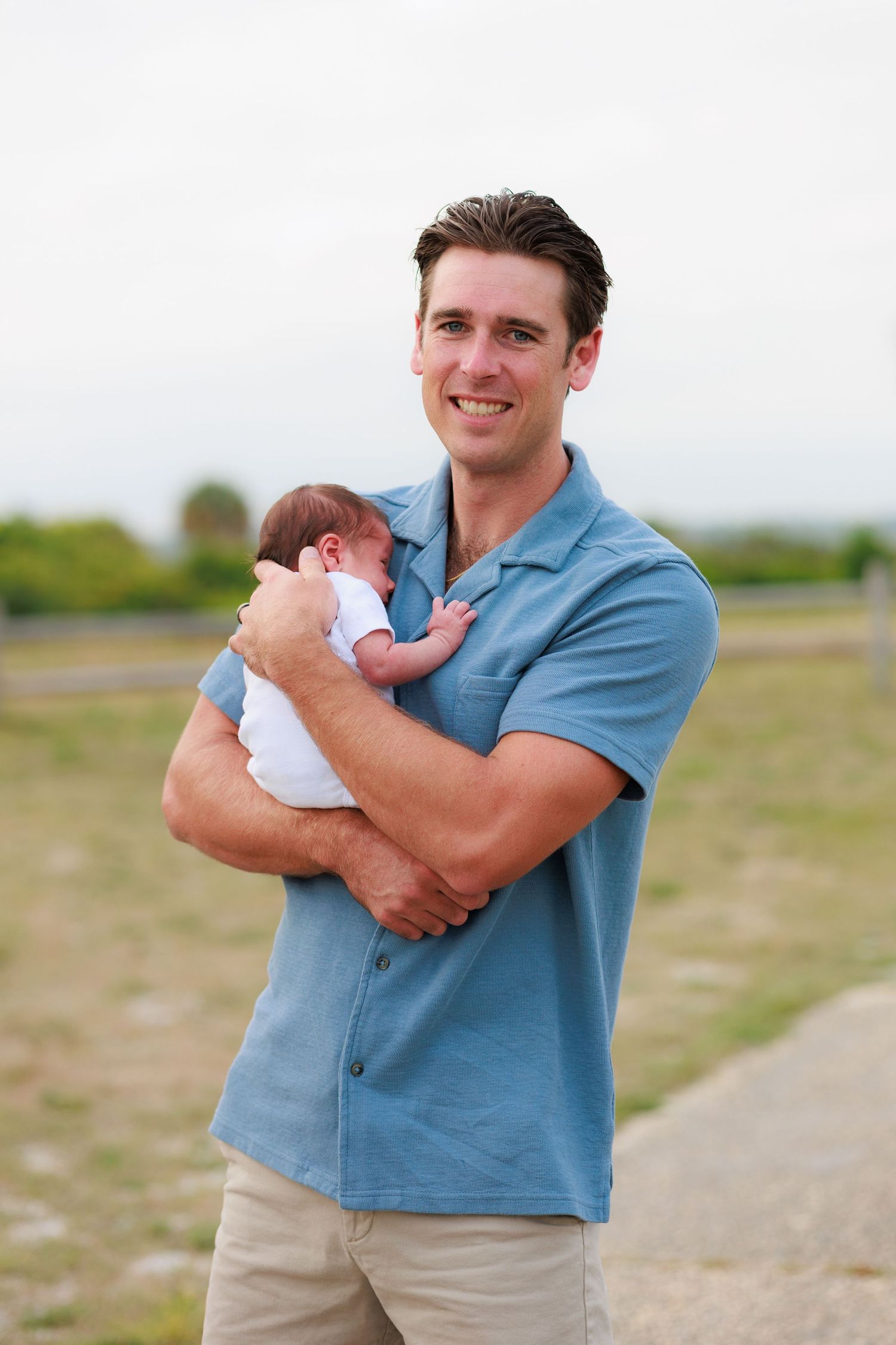 A person in a blue polo shirt holds a newborn baby while standing outdoors in a grassy field.