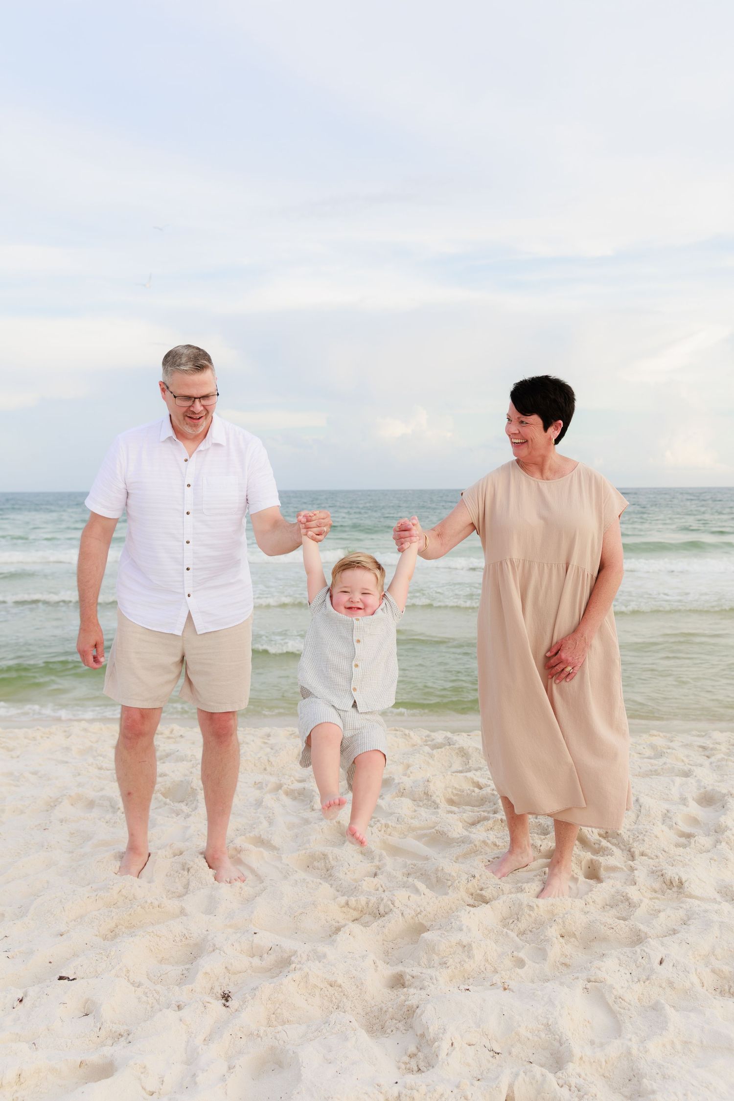 Family wearing white and beige outfits plays together on a sandy beach with ocean waves in the background.