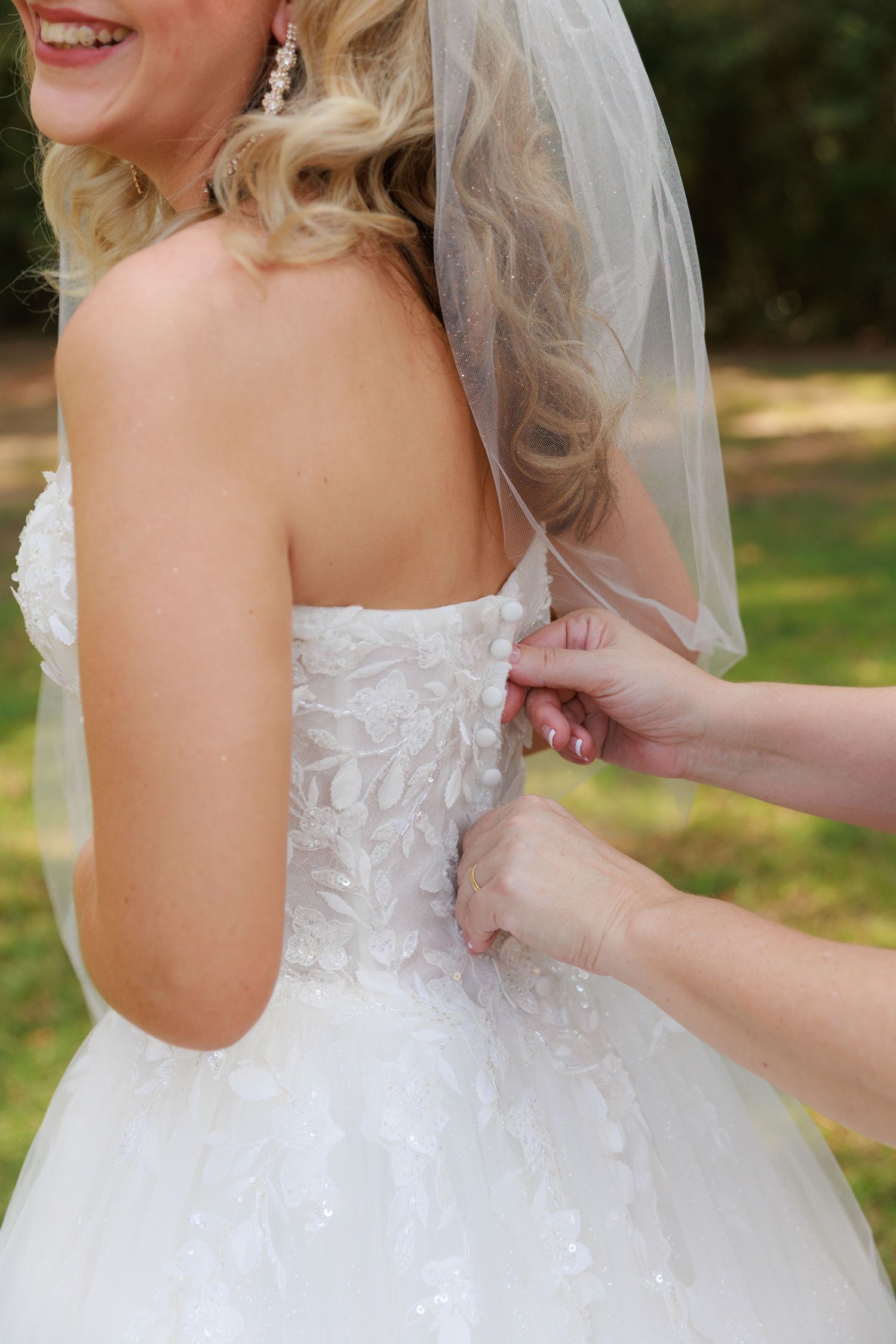 Someone helps a bride adjust the back of her white strapless wedding gown with lace details and veil.