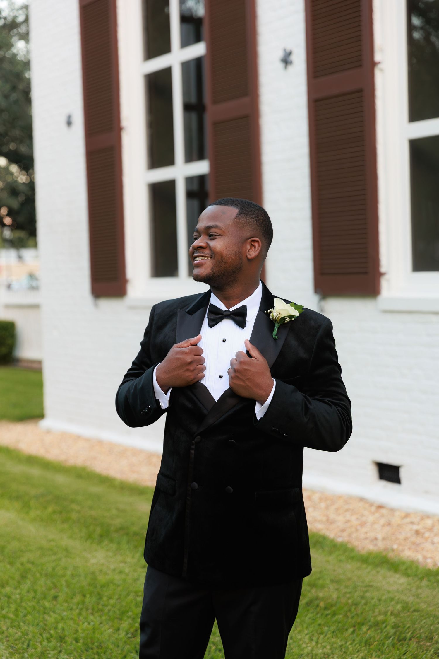 A groom in a classic black tuxedo and bowtie adjusts his lapels while smiling outside a white building with brown shutters.