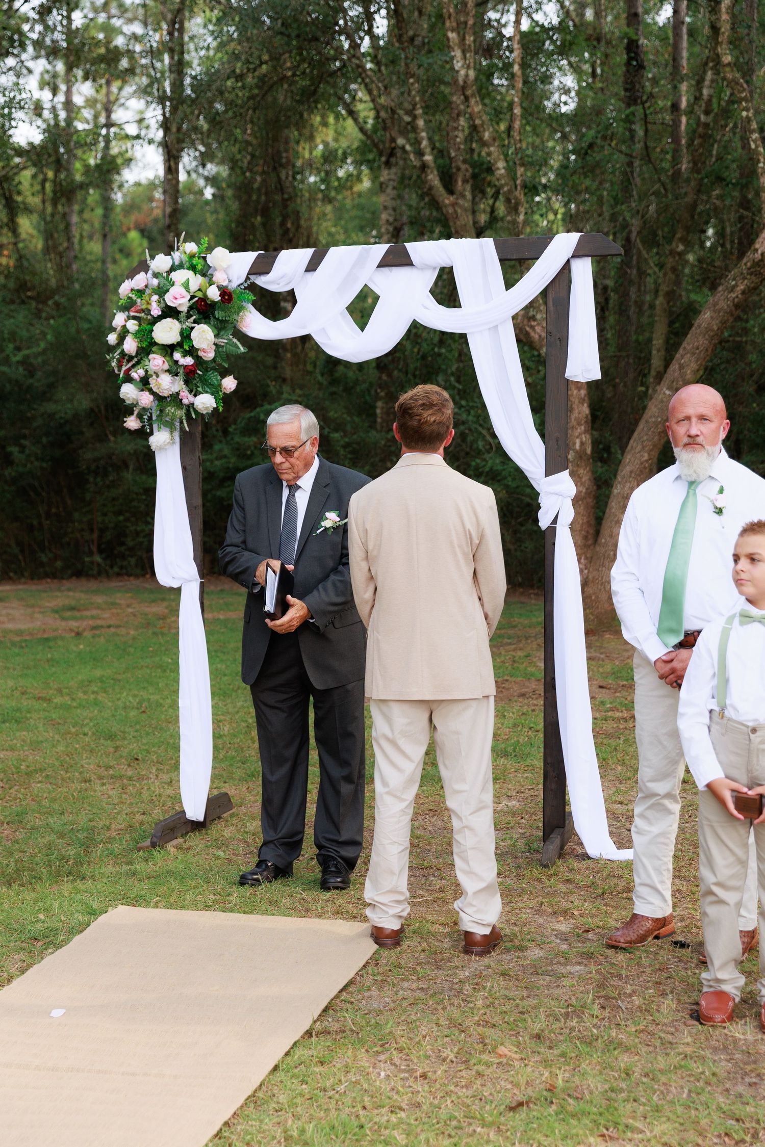 An outdoor wedding ceremony setup with a white draped arch and floral arrangement in a wooded setting.