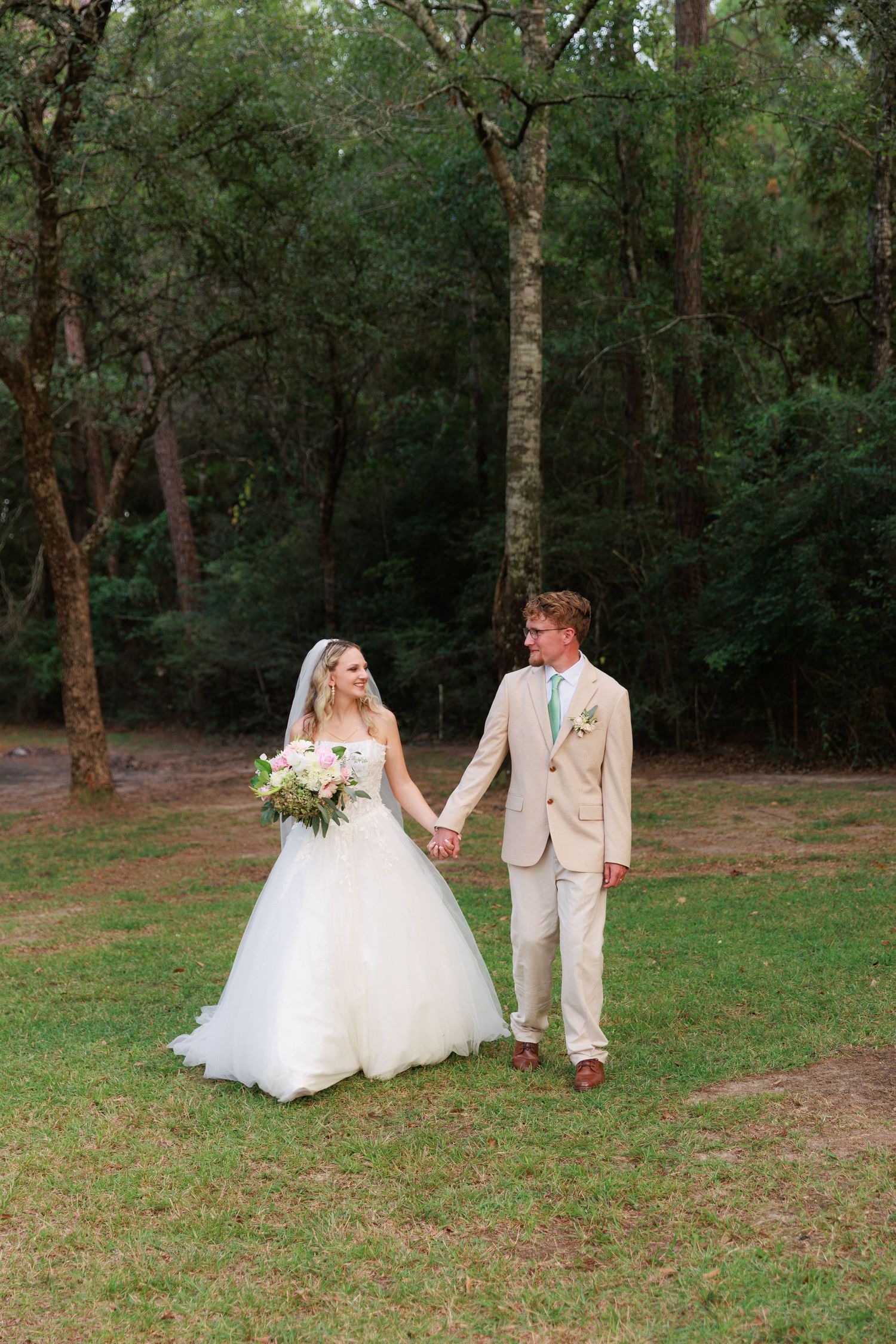 A couple in wedding attire walks through a serene wooded area with green grass and trees in the background.