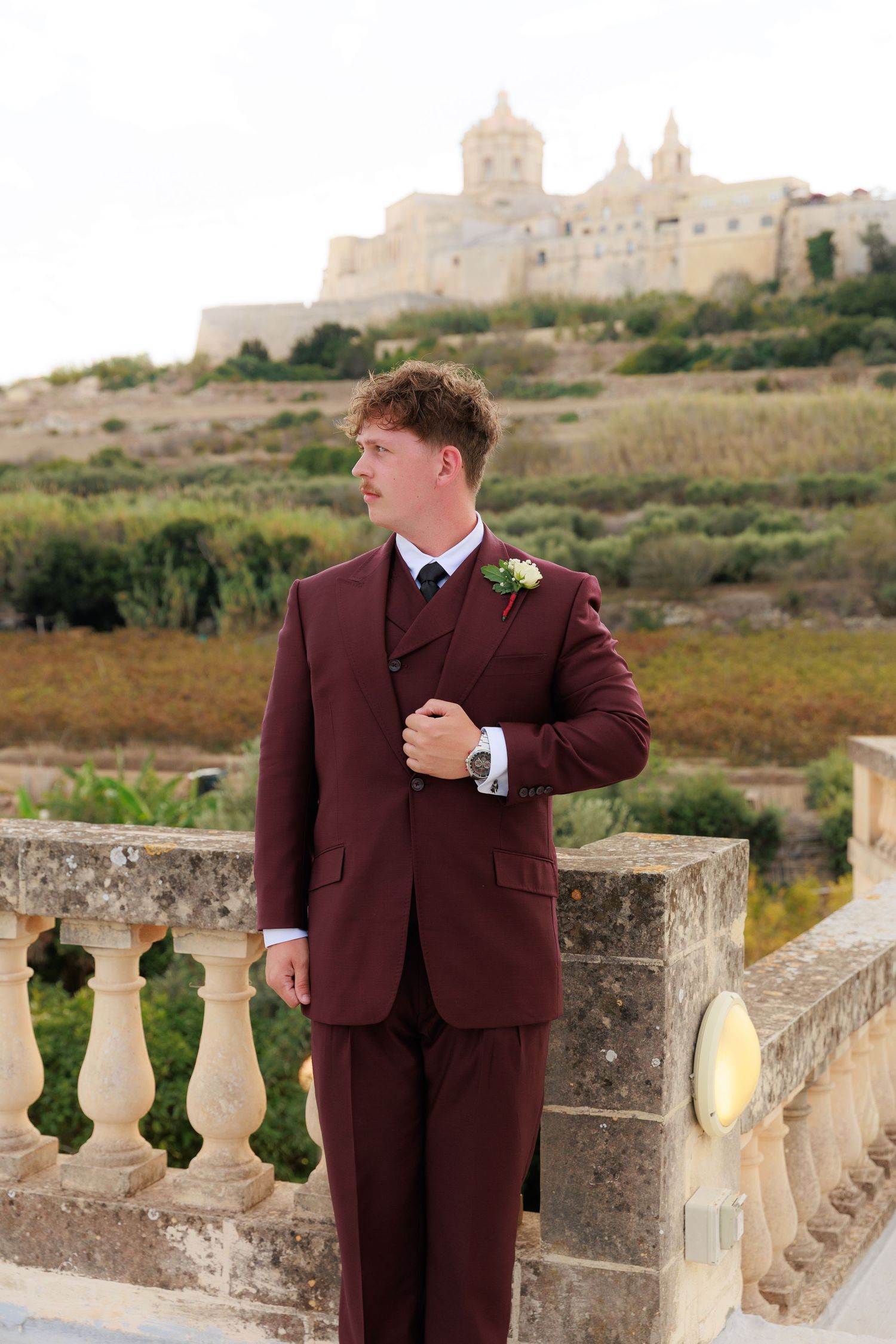 A wedding portrait featuring a burgundy suit against a historic Mediterranean castle backdrop.