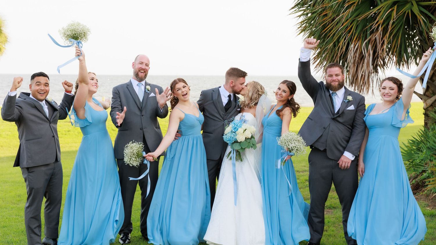 Celebratory wedding party photo with bridesmaids in blue dresses holding bouquets outdoors.