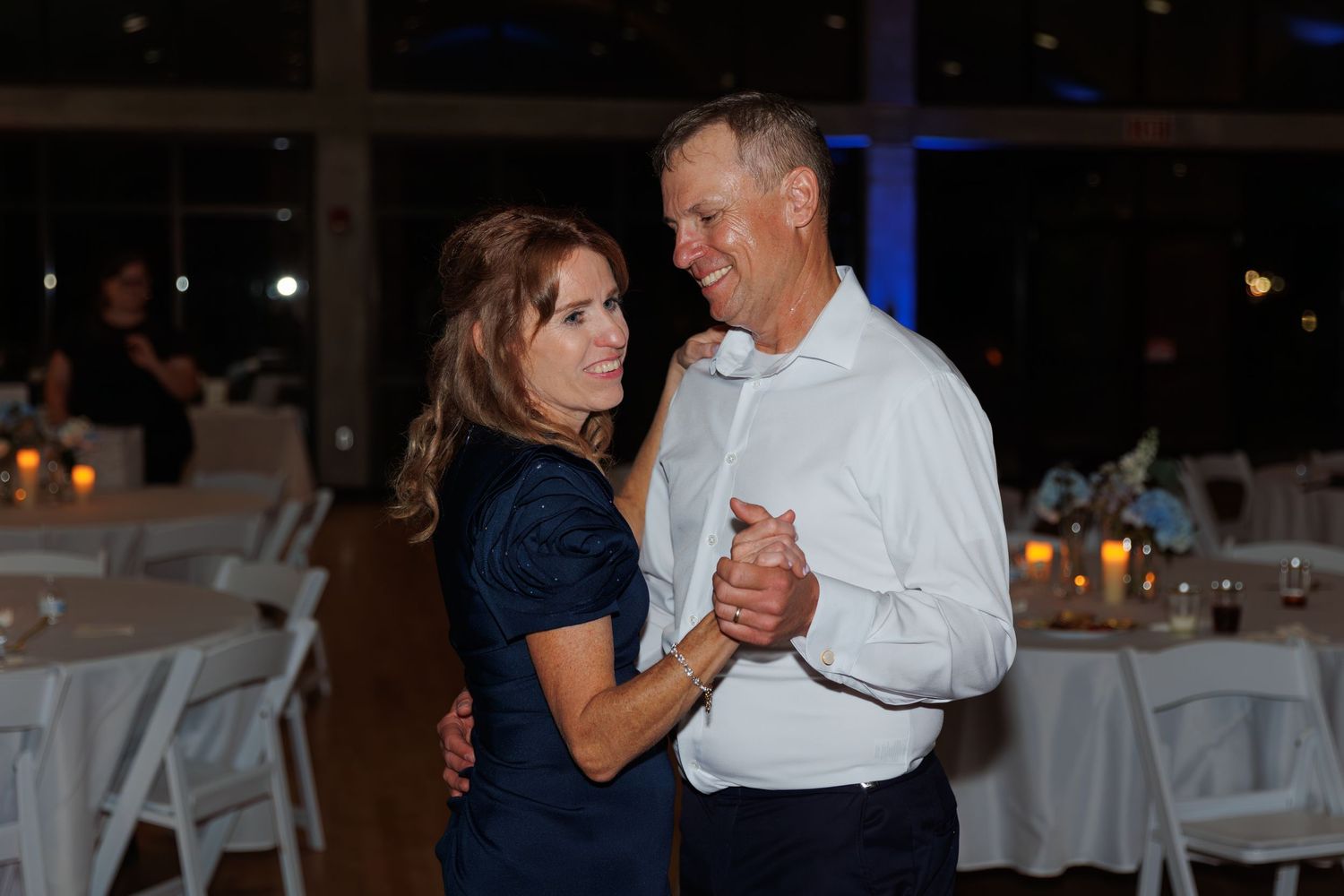 Couple dancing together at evening reception with candlelit tables in background.