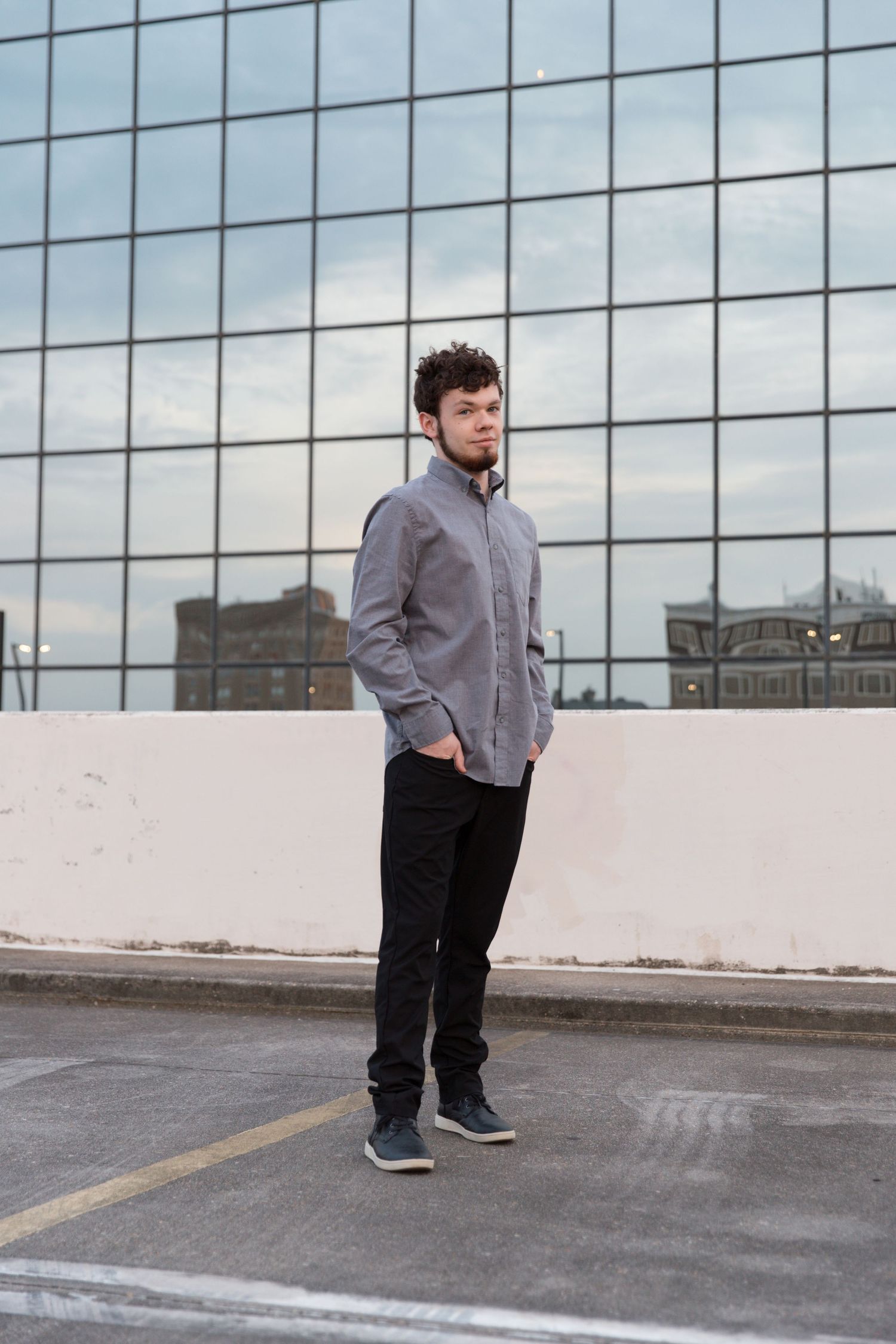 Person wearing grey button-down and black pants standing in front of modern glass building.