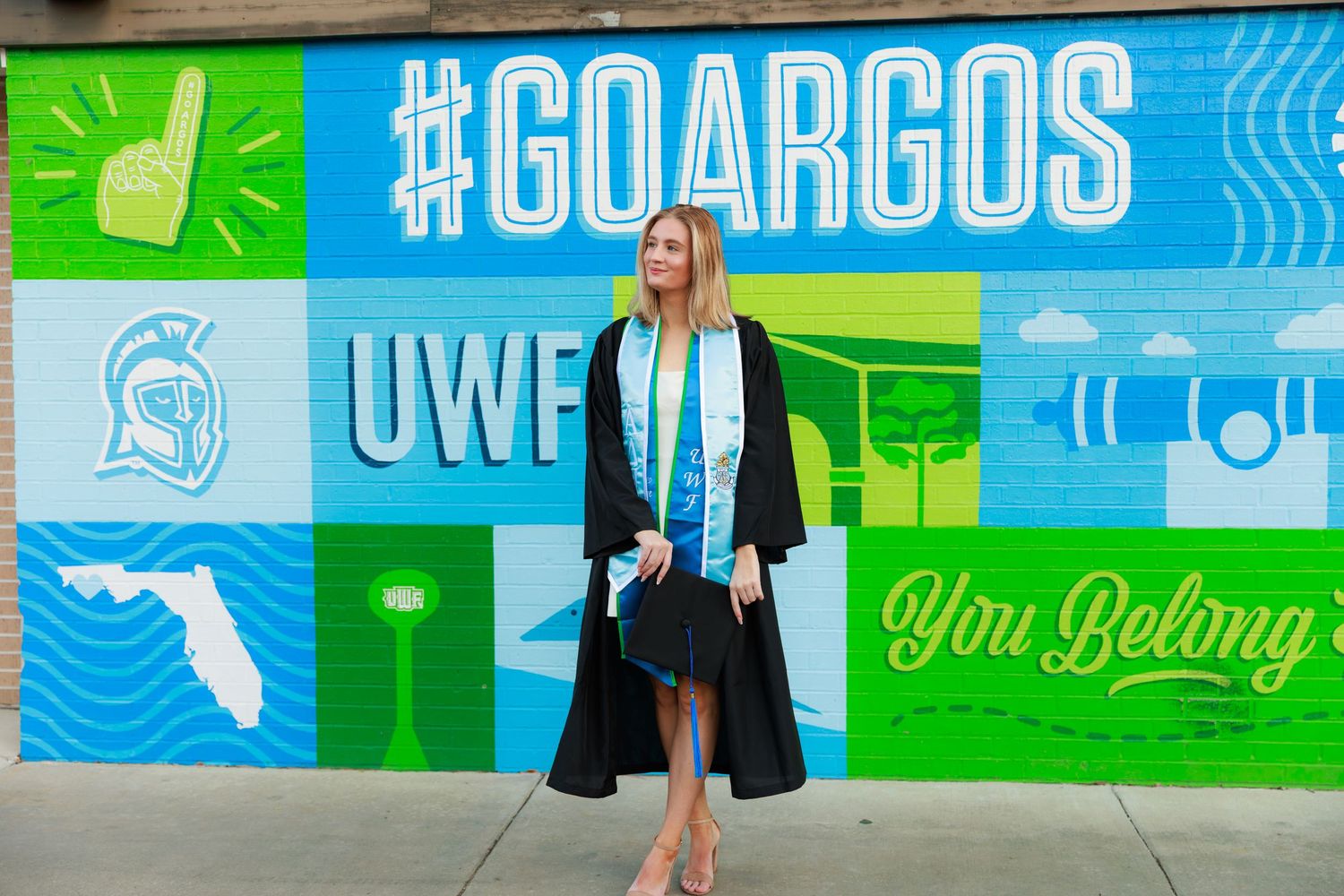 Graduate in cap and gown poses in front of bright blue and green UWF #GOARGOS mural wall.