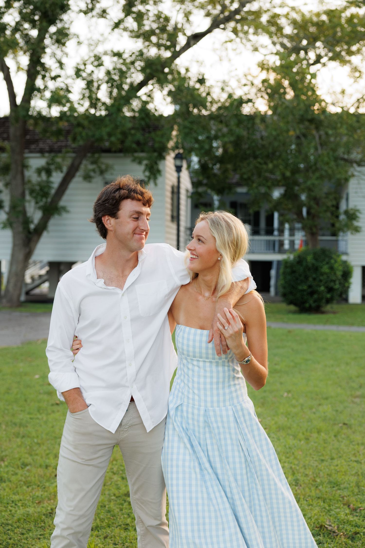 A couple in summer attire smiles together on a green lawn with a white house and trees in the background.