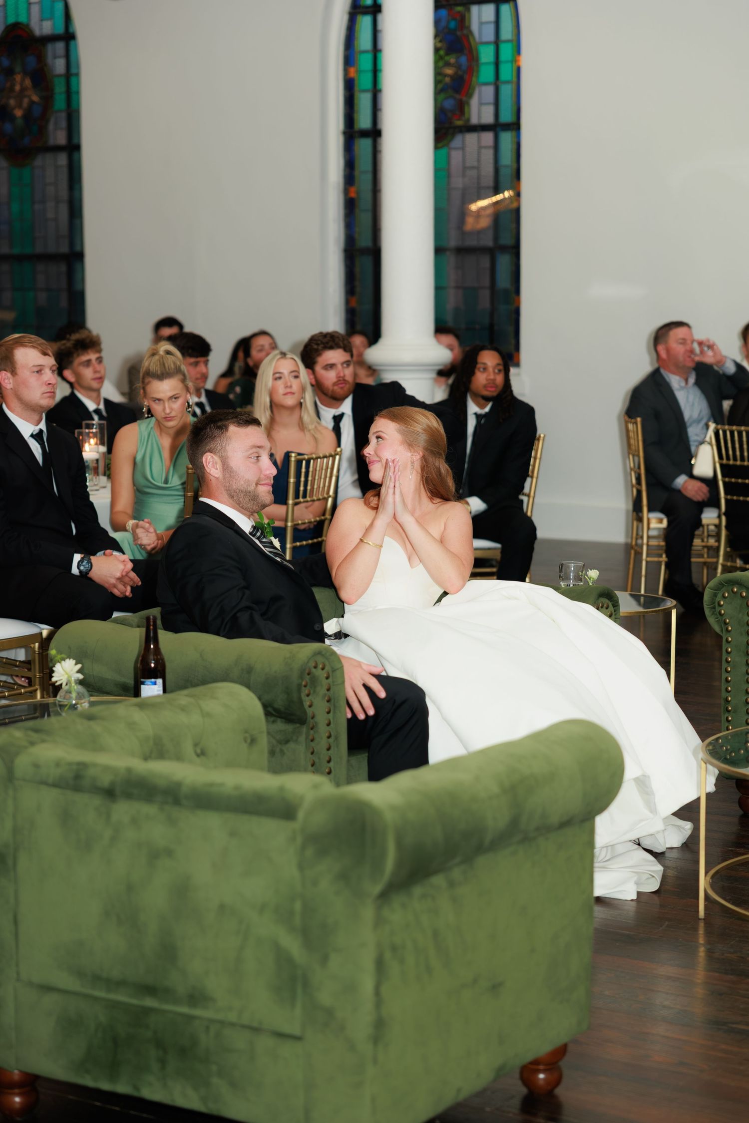 Bride with hands on cheeks sitting beside groom on green velvet sofa during wedding reception in elegant church.